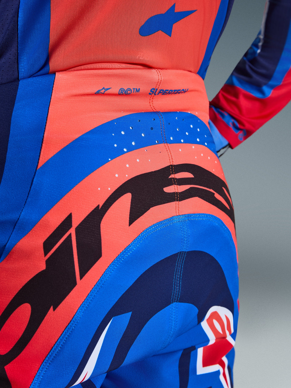 Close-up of Alpinestars 2026 Supertech Vista Pants, Motocross Pants, Hot Coral and Cobalt Blue, featuring a large black Alpinestars graphic on the seat, laser-cut ventilation holes, and 'SUPERTECH' branding on the back waist, professional-grade racing apparel.