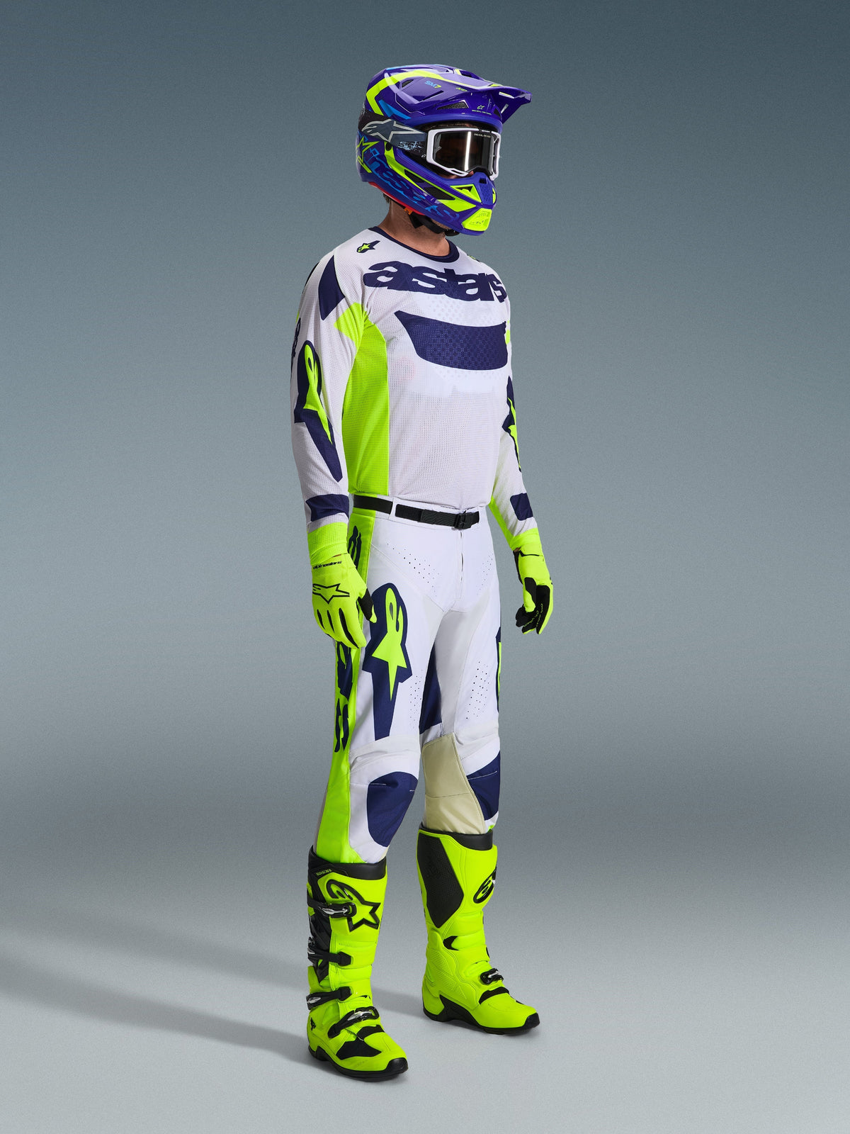 Motocross rider wearing the Alpinestars 2026 Racer Air Riway Gear Set in Light Gray, Yellow Fluo, and Blue. The set includes a long-sleeve mesh jersey, matching racing pants, bright neon yellow gloves and boots, and a purple and blue helmet with goggles, designed for ventilated off-road performance.