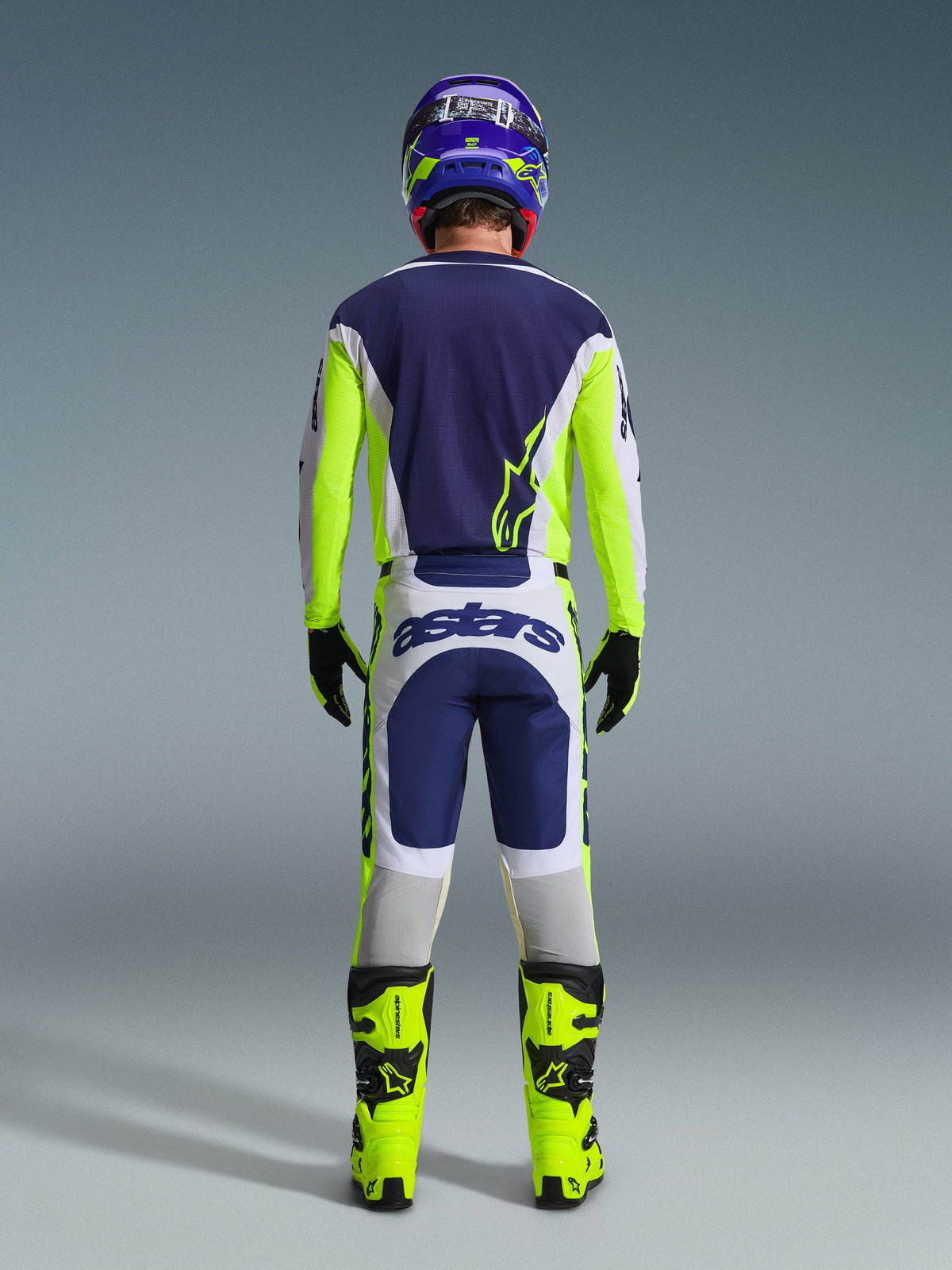 Motocross rider from behind wearing the Alpinestars 2026 Racer Air Riway Gear Set, Blue jersey with fluo yellow and light gray accents, white and blue pants with fluo yellow side panels, fluo yellow boots with black details, matching blue and yellow helmet, professional gear for warm-weather off-road riding