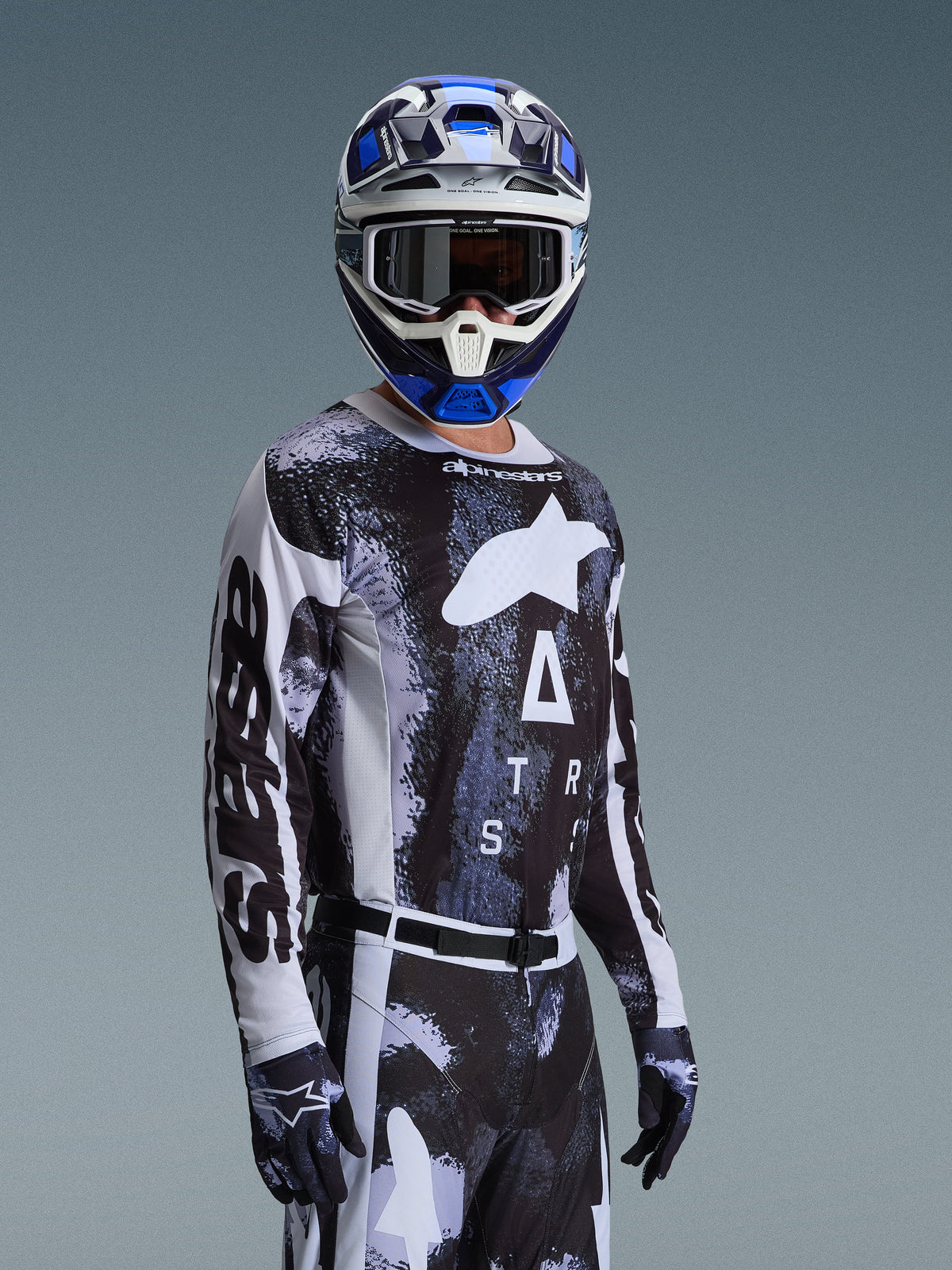 Motocross rider wearing Alpinestars 2026 Racer Lahnd Gear Set, black and white jersey with marbled blue and grey graphics, matching pants and gloves, blue and white helmet with goggles, performance gear for off-road racing