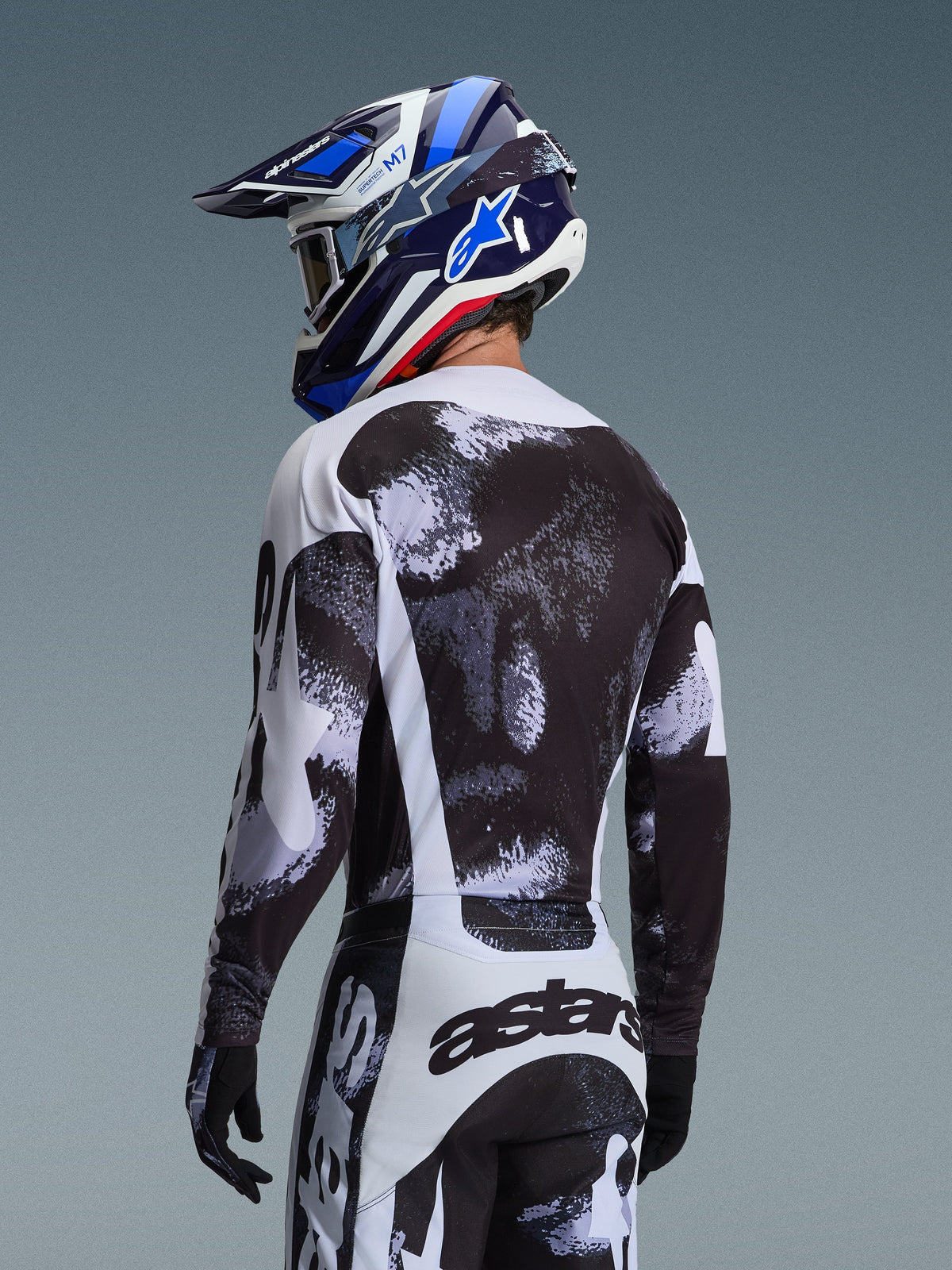 Alpinestars 2026 Racer Lahnd Jersey in Iron Camo, rear view of a motocross rider in a black, white, and grey abstract pattern gear set, including matching pants, black gloves, and a blue racing helmet with goggles.