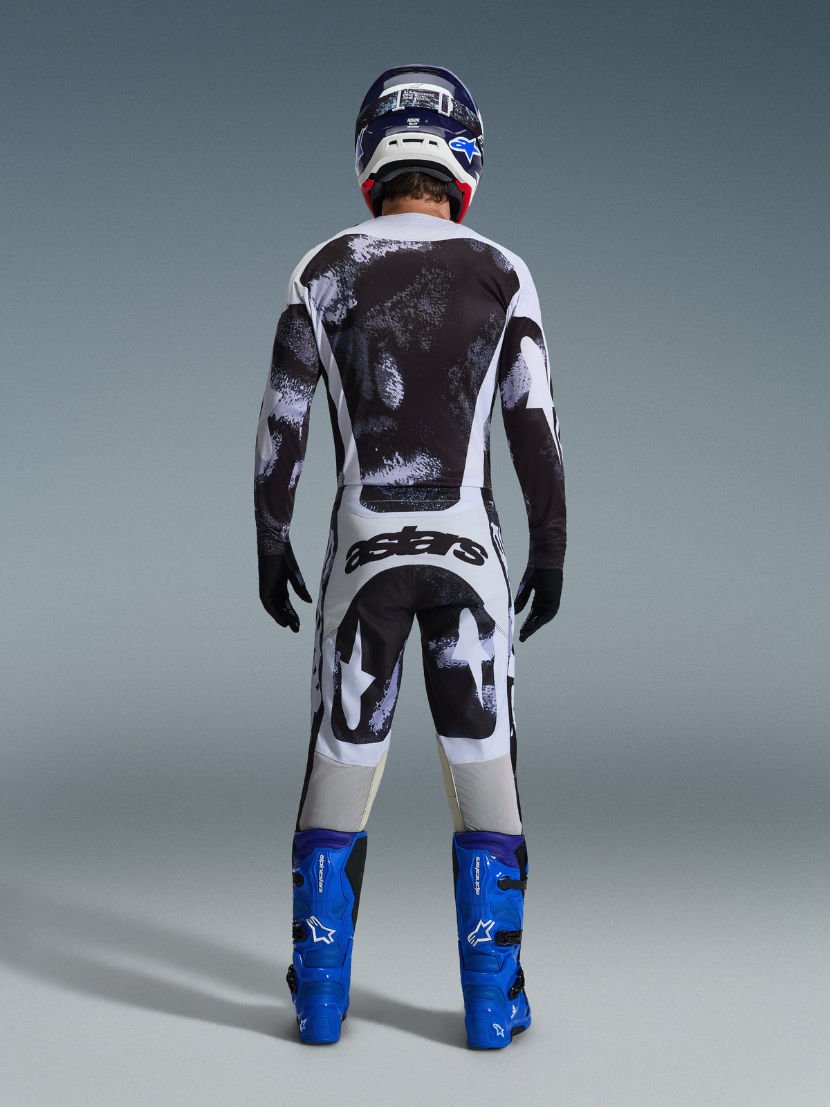 Motocross rider from the rear wearing the Alpinestars 2026 Racer Lahnd Gear Set, black and white abstract patterned jersey and pants, 'astars' branding on the seat, paired with blue boots and a matching helmet, professional off-road racing apparel.