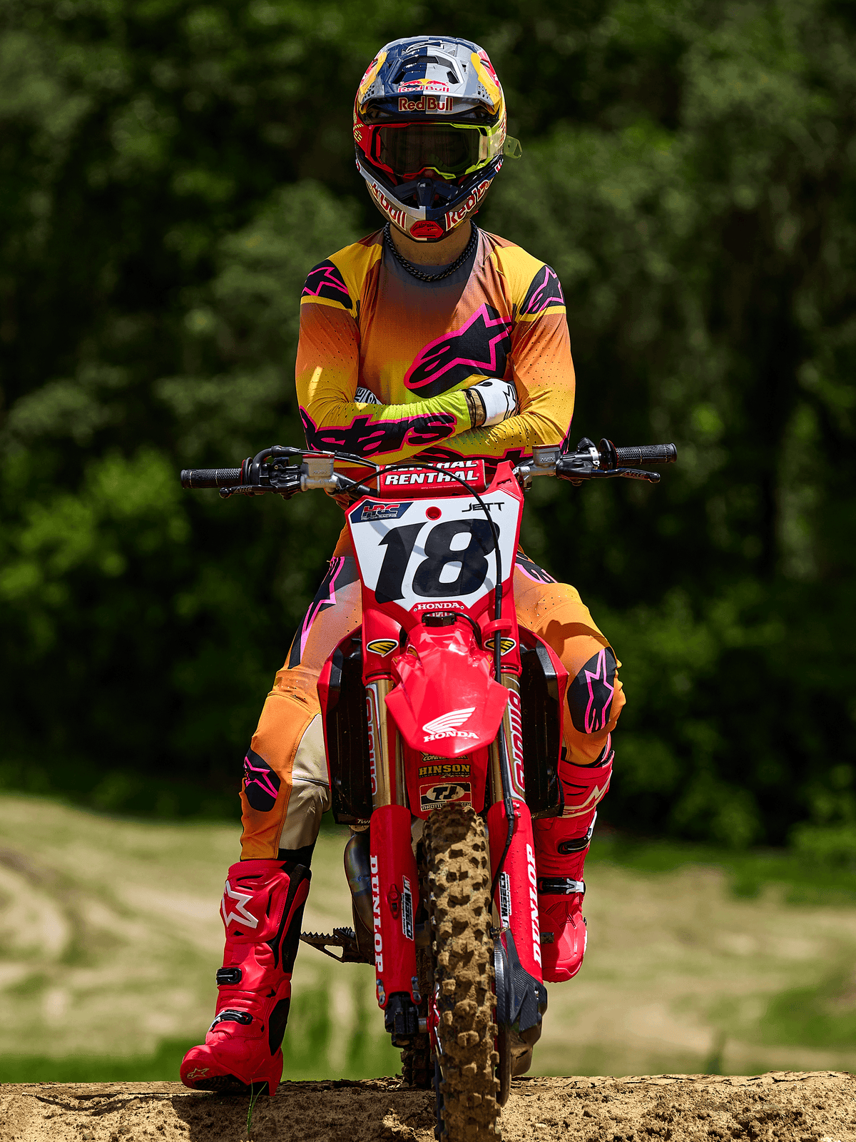 Motocross rider sitting on a red Honda dirt bike with racing number 18, wearing Alpinestars 2025 Supertech Lipan Jersey and matching pants in Yellow and Pink, bright pink Alpinestars boots, Red Bull helmet, professional racing gear for off-road performance.