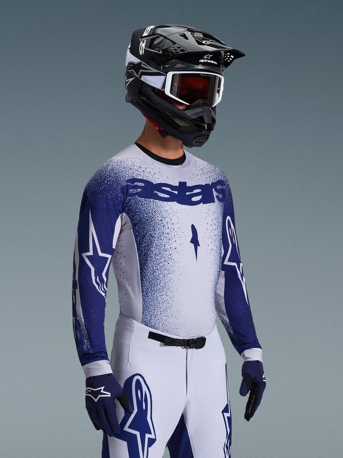 Motocross rider wearing the 2026 Supertech Scenz Jersey in Light Gray and Dark Blue, paired with matching white and blue racing pants, blue gloves, and a black helmet with white goggles. The jersey features a stippled blue gradient effect on the chest and sleeves, professional athletic fit, and bold 'astars' branding.
