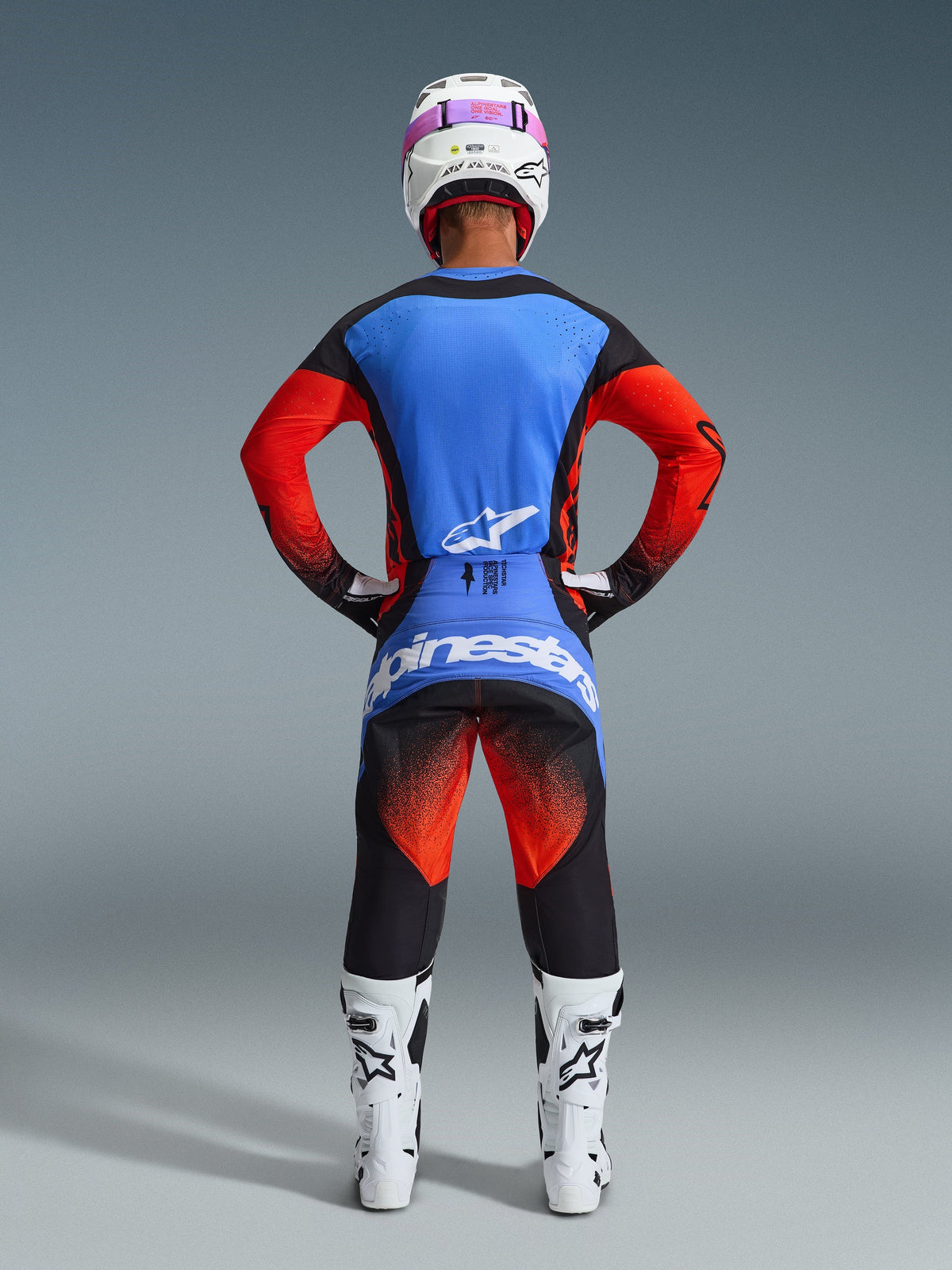 Motocross rider from behind wearing complete racing gear, 2026 Techstar Knif Jersey with blue back and orange sleeves, black pants with orange and blue accents, white helmet with pink goggle strap, white and black boots, coordinated outfit for off-road racing