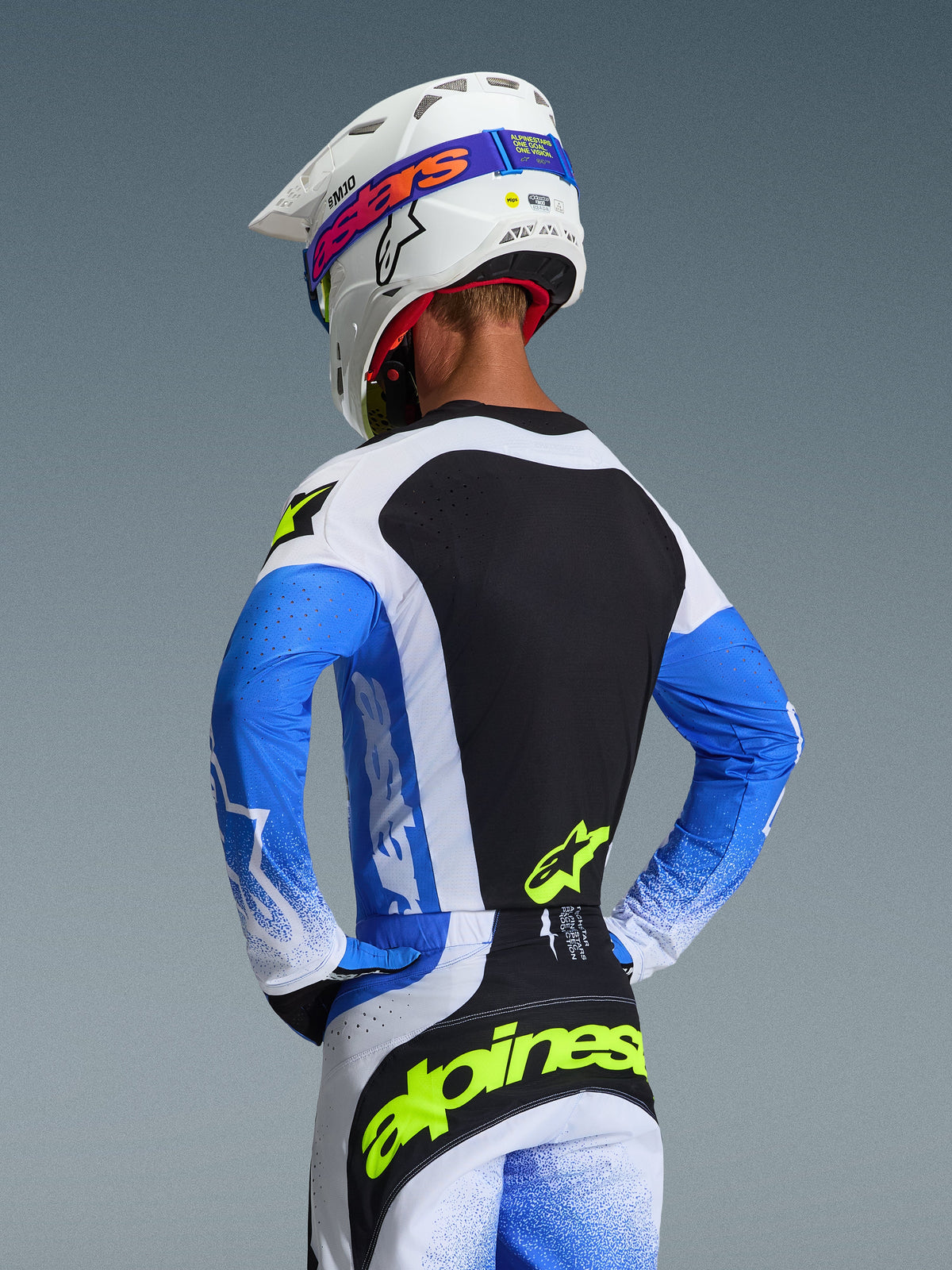Rear view of a motocross rider wearing the Alpinestars 2026 Techstar Knif Jersey in Ucla Blue, Black, and Yellow Fluo, paired with matching pants and a white Supertech M10 helmet, athletic race fit gear for off-road performance.