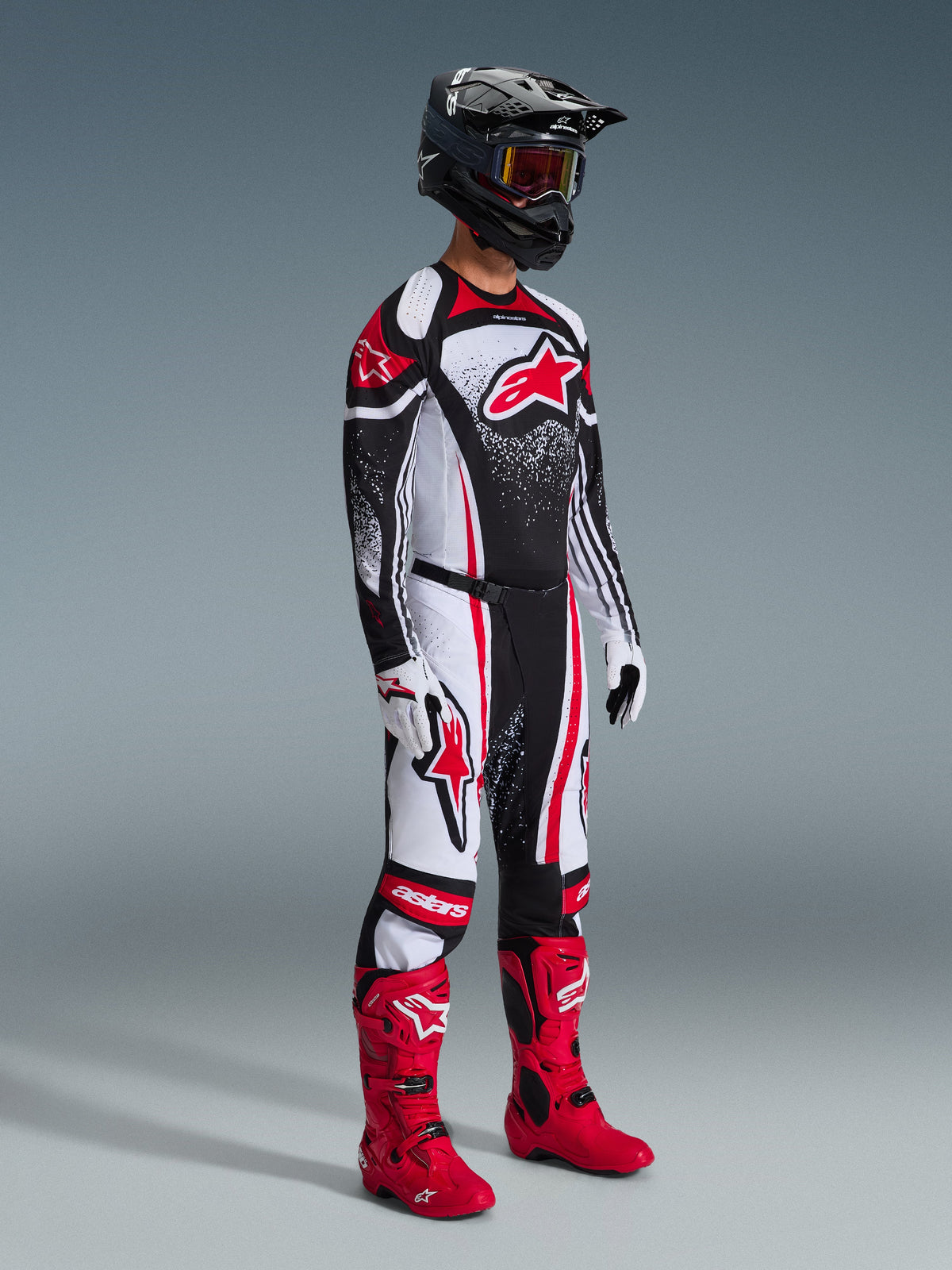 Motocross rider wearing the Alpinestars 2026 Techstar Nomur Gear Set, black jersey with red and white graphic patterns, matching motocross pants, bright red boots, black helmet with goggles, coordinated off-road racing outfit