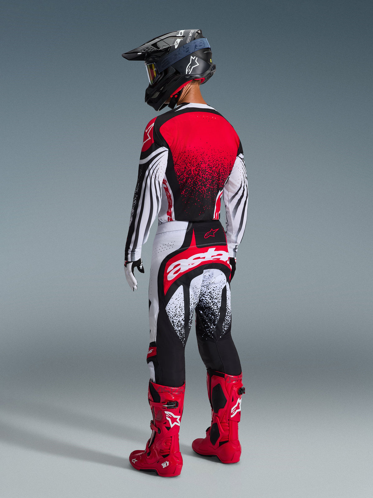 Motocross rider from the rear view wearing the Alpinestars 2026 Techstar Nomur Gear Set, Red White and Black jersey with dotted gradient, matching pants with bold 'astars' branding, black helmet, and red motocross boots, professional off-road racing kit