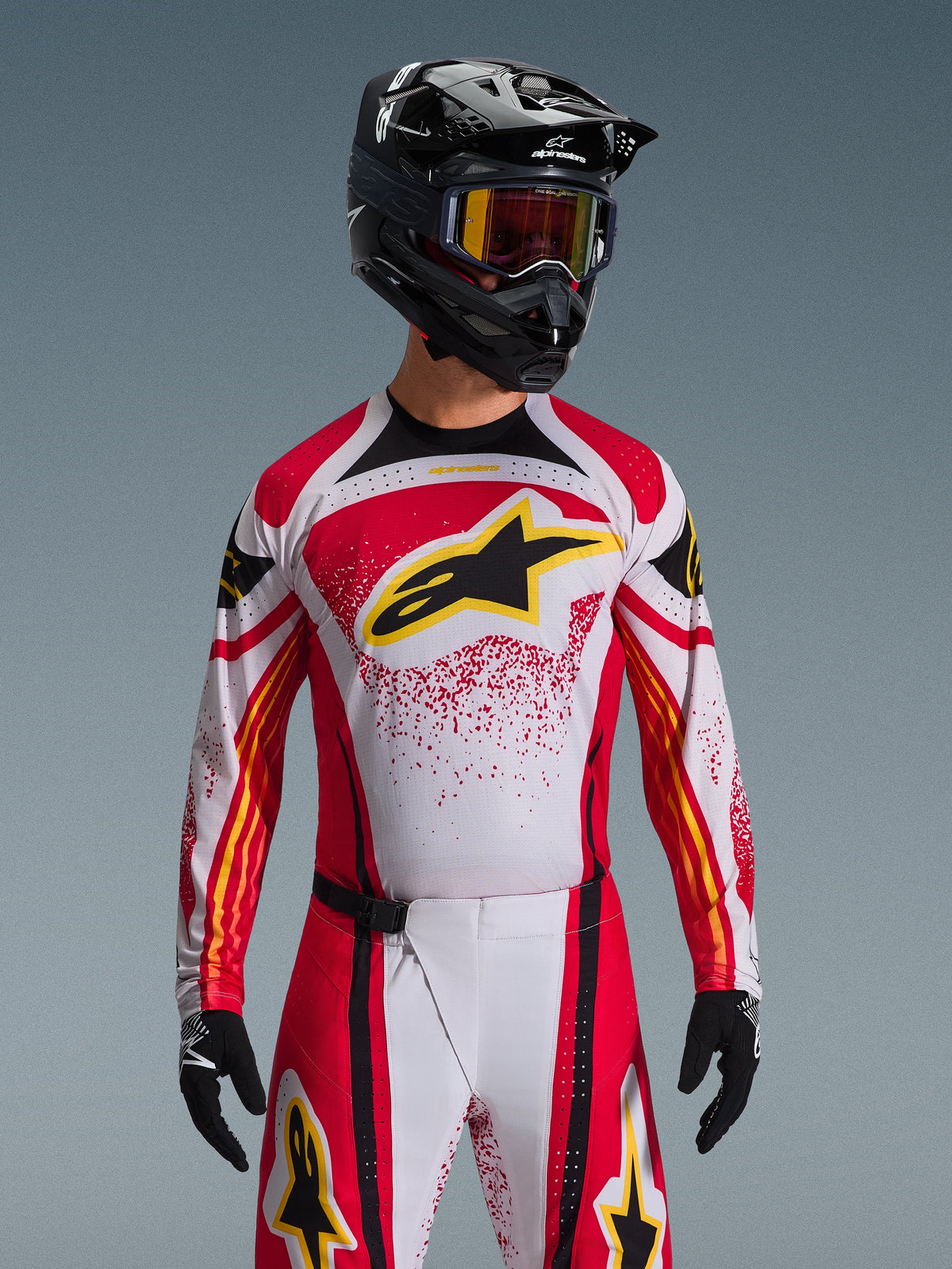 Alpinestars 2026 Techstar Nomur Gear Set, Motocross rider wearing red, white, and black jersey with yellow logo, matching pants with speckled pattern, black helmet and goggles, black gloves, complete racing gear for off-road competition