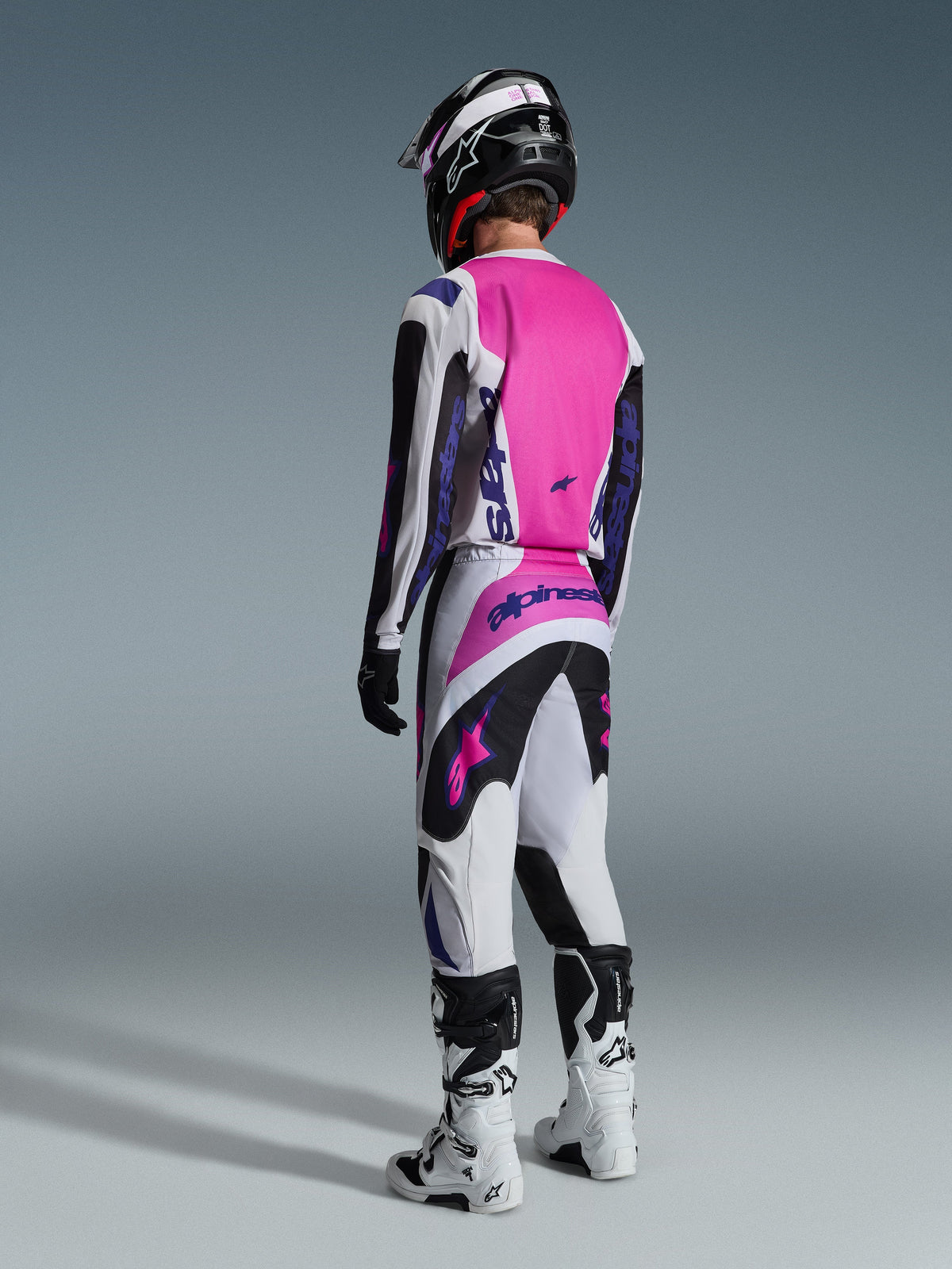 Motocross rider from behind wearing the Alpinestars 2026 Fluid Grid Jersey and matching pants, light gray and black with a vibrant pink back panel and purple branding, complete with a black helmet and white boots.