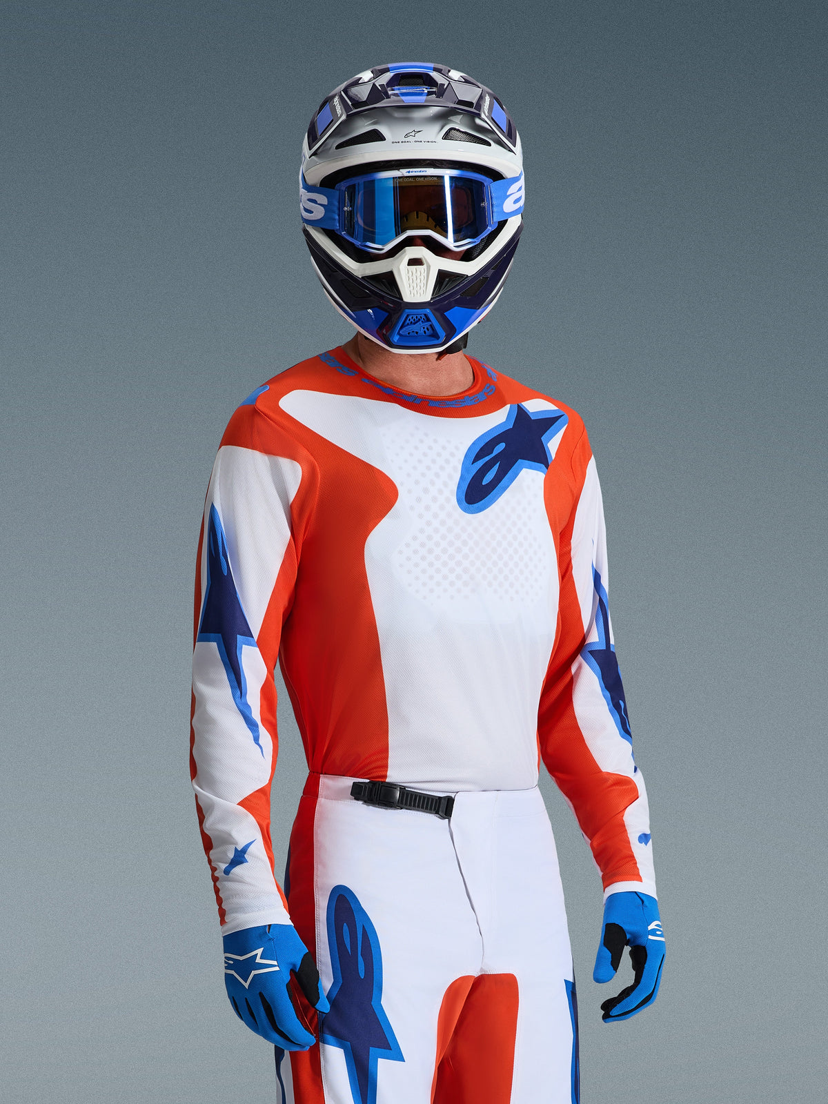 Motocross rider wearing the Alpinestars 2026 Fluid Grid Gear Set, orange and white jersey with blue star logos, white and orange pants with blue accents, blue gloves, blue and white helmet with blue goggles, coordinated athletic racing gear for off-road performance