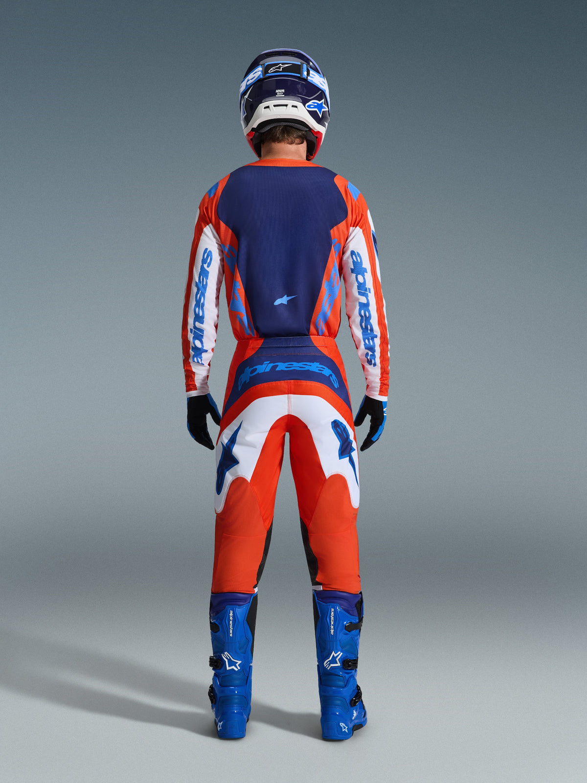 Alpinestars 2026 Fluid Grid Gear Set, Motocross rider from the back wearing a complete racing outfit in Orange and UCLA Blue, long-sleeve jersey with navy blue back and white sleeves, matching orange pants with blue graphics, blue boots, and coordinated helmet, professional off-road racing gear.