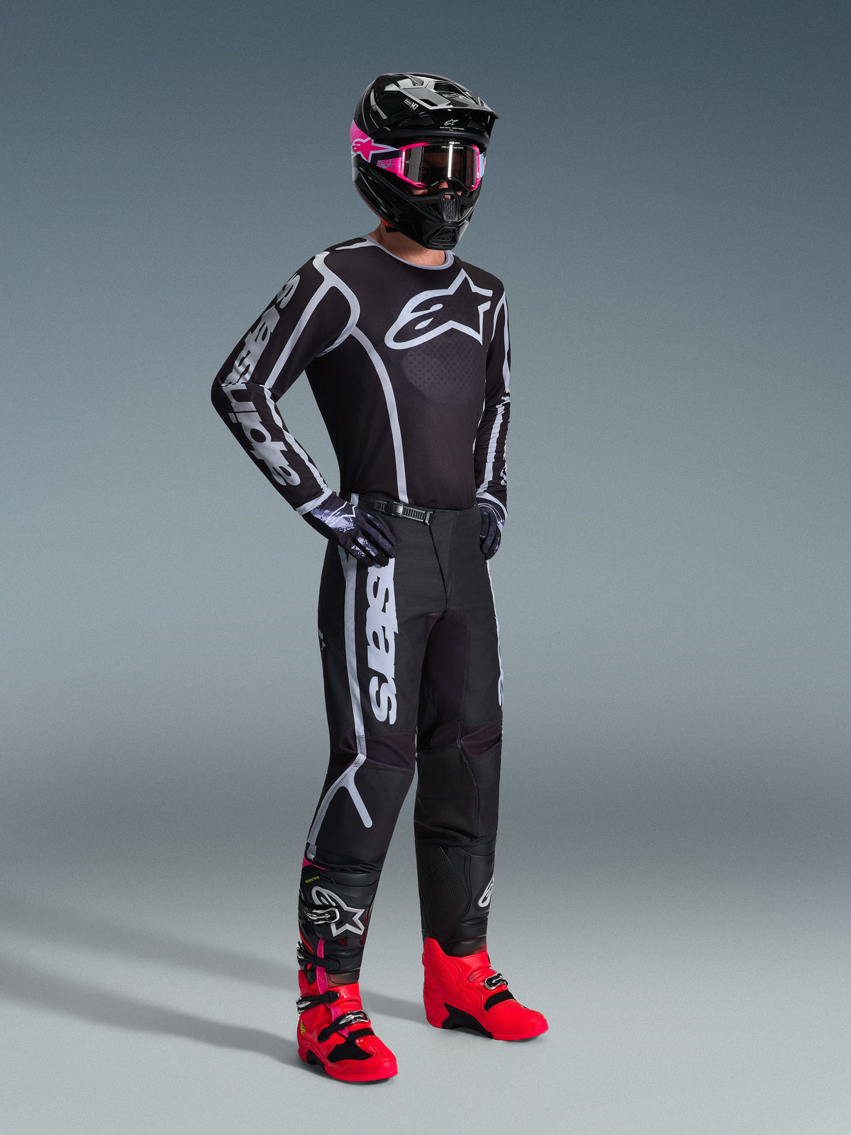 Alpinestars 2026 Fluid Apex Gear Set, motocross rider wearing a black jersey and pants with white and grey graphic accents, black helmet with pink goggles, and bright pink racing boots, coordinated professional motocross outfit