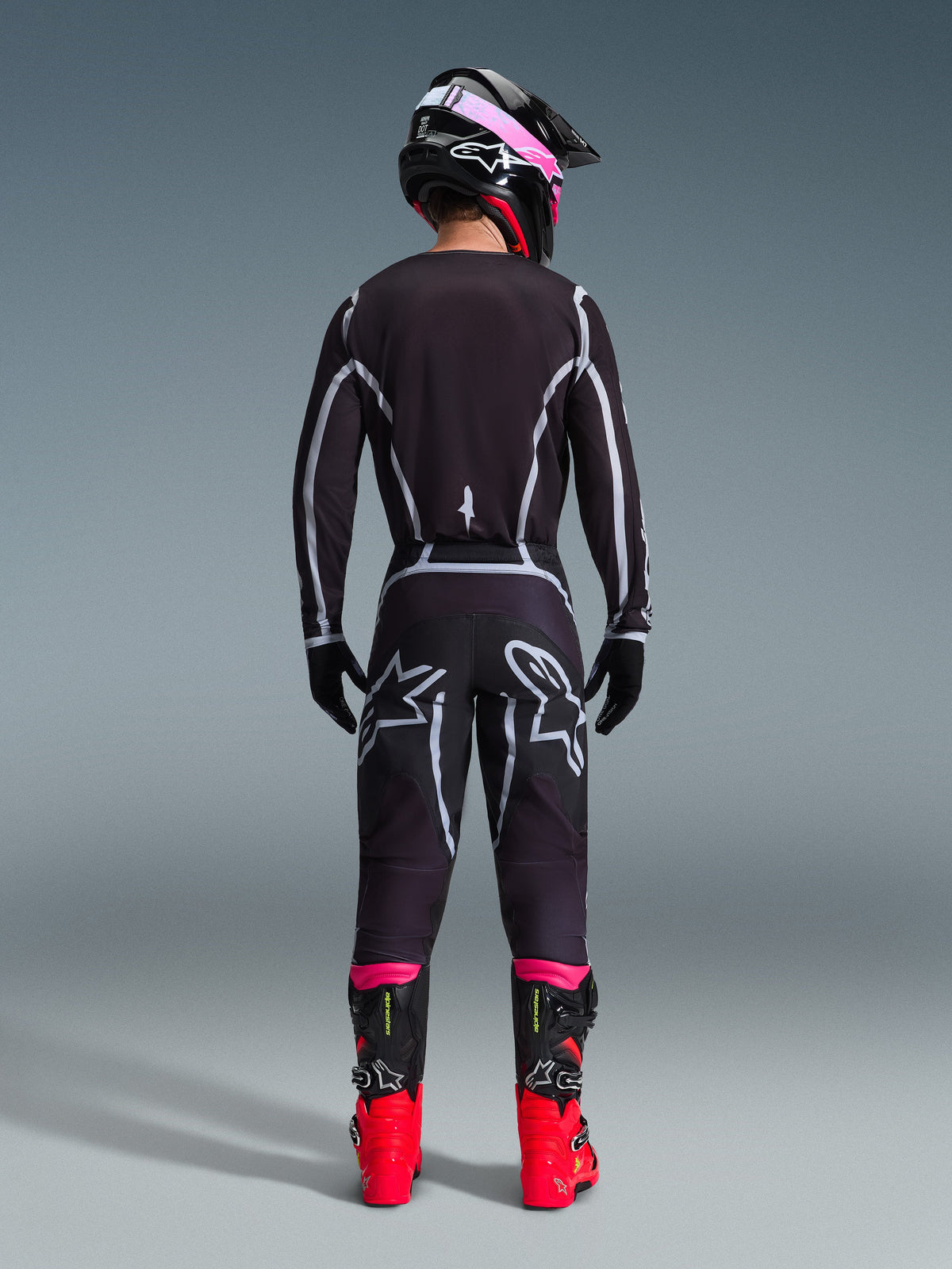 Motocross rider seen from behind wearing the Alpinestars 2026 Fluid Apex Gear Set, black jersey and pants with grey geometric lines and star logos, black helmet with pink accents, bright red and black boots, coordinated motocross racing gear