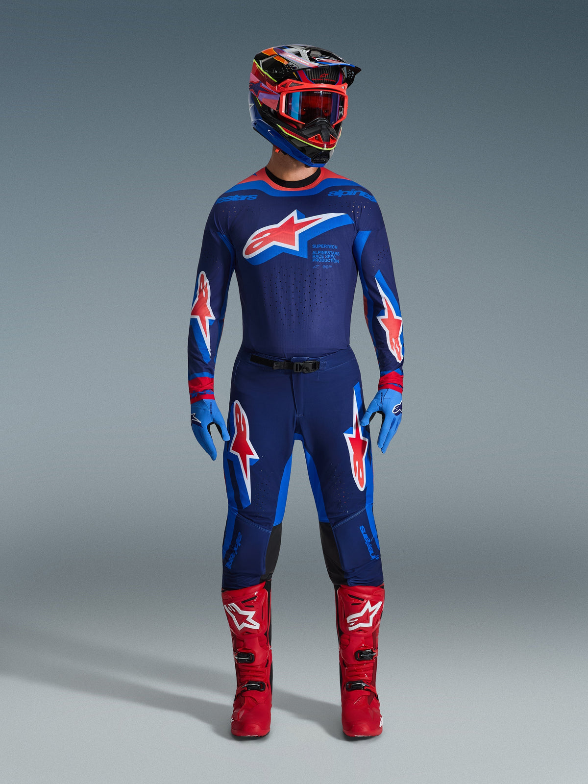 Motocross rider wearing the Alpinestars 2026 Supertech Vista Jersey in Dark Navy, Hot Coral, and Cobalt, paired with matching racing pants, bright red boots, and a coordinated helmet with red goggles, complete professional gear set for off-road performance.