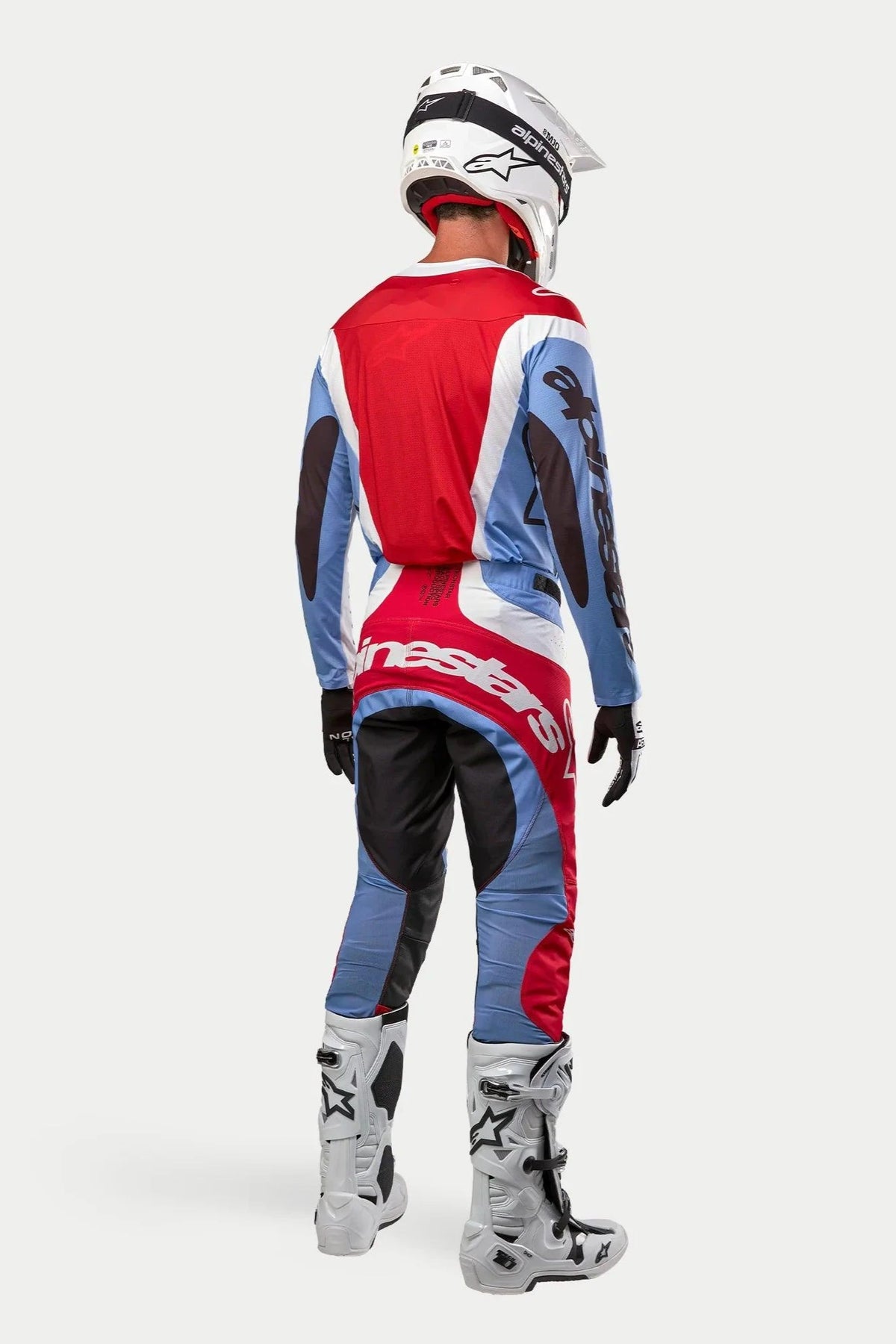 Motocross rider seen from the back wearing complete racing gear, 2024 Techstar Ocuri Jersey in Light Blue, Mars Red, and White, matching pants with bold branding, white and black boots, white helmet, coordinated outfit for off-road racing