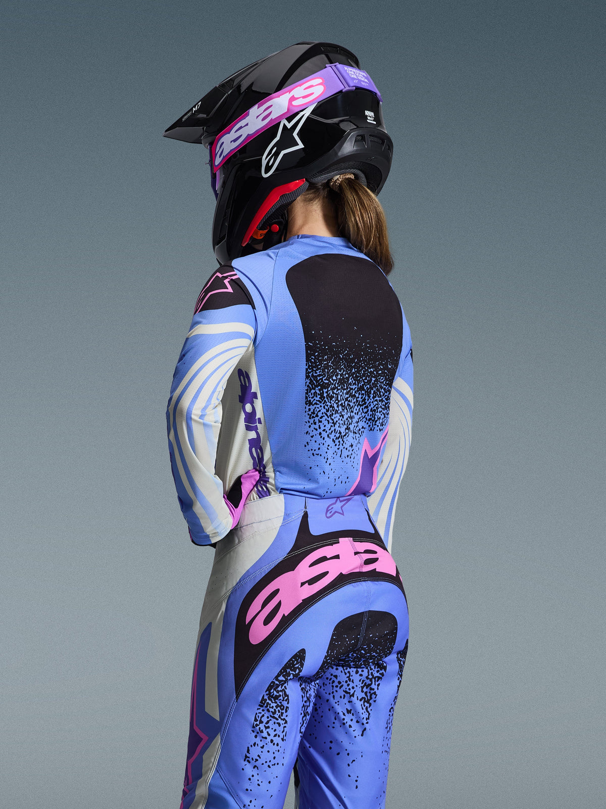 Rear view of a motocross rider wearing the Alpinestars 2026 Stella Techstar Nomur Gear Set, Light Blue with Pink and Black accents, matching jersey and pants, black helmet with pink goggles, ergonomic women's fit for off-road racing.