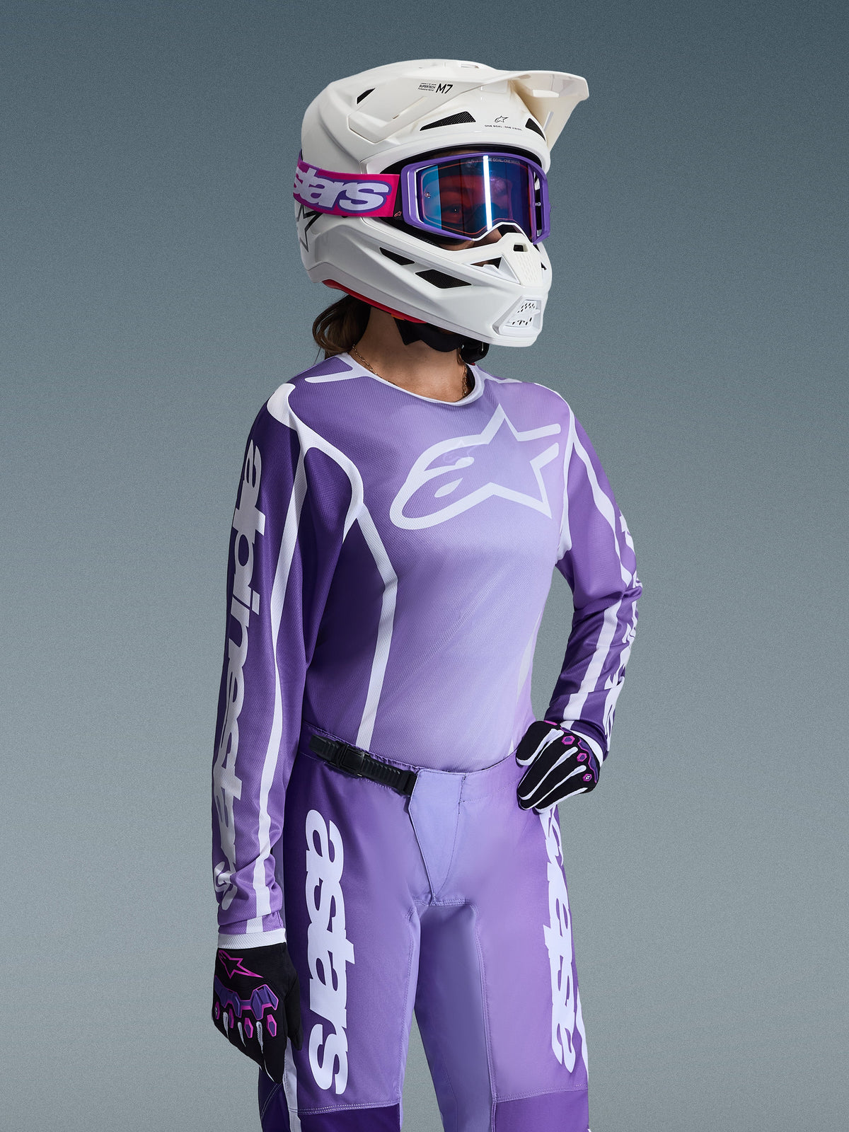 Motocross rider wearing Alpinestars 2026 Woman Stella Fluid Apex Gear Set, Purple and white jersey with Alpinestars logos, matching purple pants, white helmet with purple goggles, and black/purple gloves, professional off-road racing gear