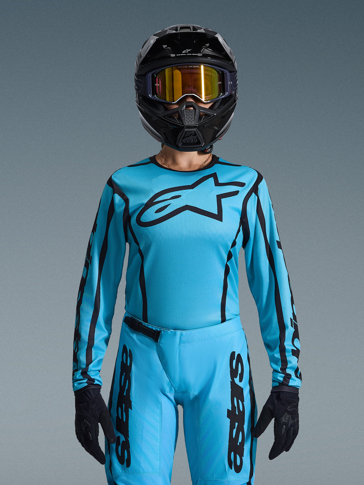 Motocross rider wearing Alpinestars 2026 Woman Stella Fluid Apex Gear Set, light blue jersey with black outlines and logo, matching pants, black helmet with iridescent gold goggles, and black gloves, coordinated women's motocross racing outfit