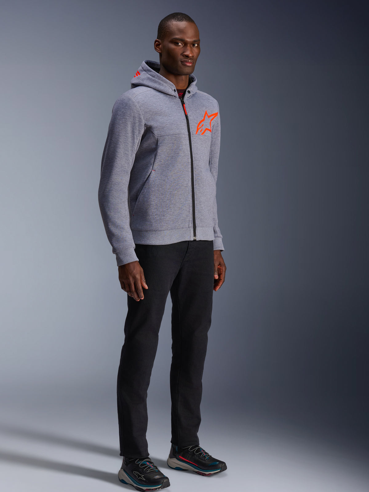 Alpinestars Chrome V2 Sport Hoodie, Gray Heather and Red Fluo, worn by a male model with black pants and dark athletic shoes, featuring a bright orange star logo on the chest and hood, casual urban riding apparel