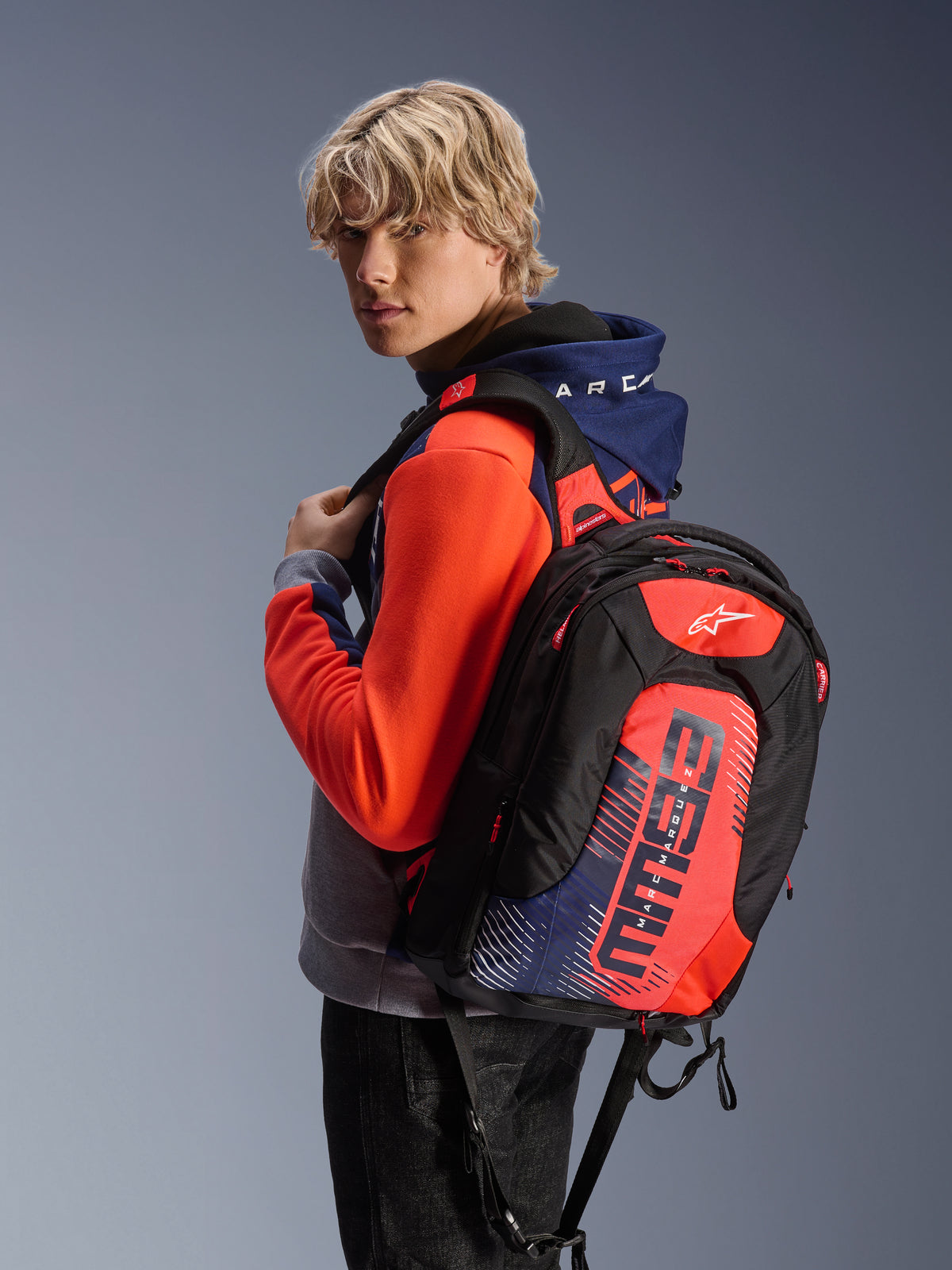 Male model wearing the Alpinestars MM93 Chrome V2 Sport Hoodie in Blue Navy Gray Mel and Red Fluo, paired with a matching MM93 branded backpack, casual urban motorcycle apparel with a modern athletic silhouette.