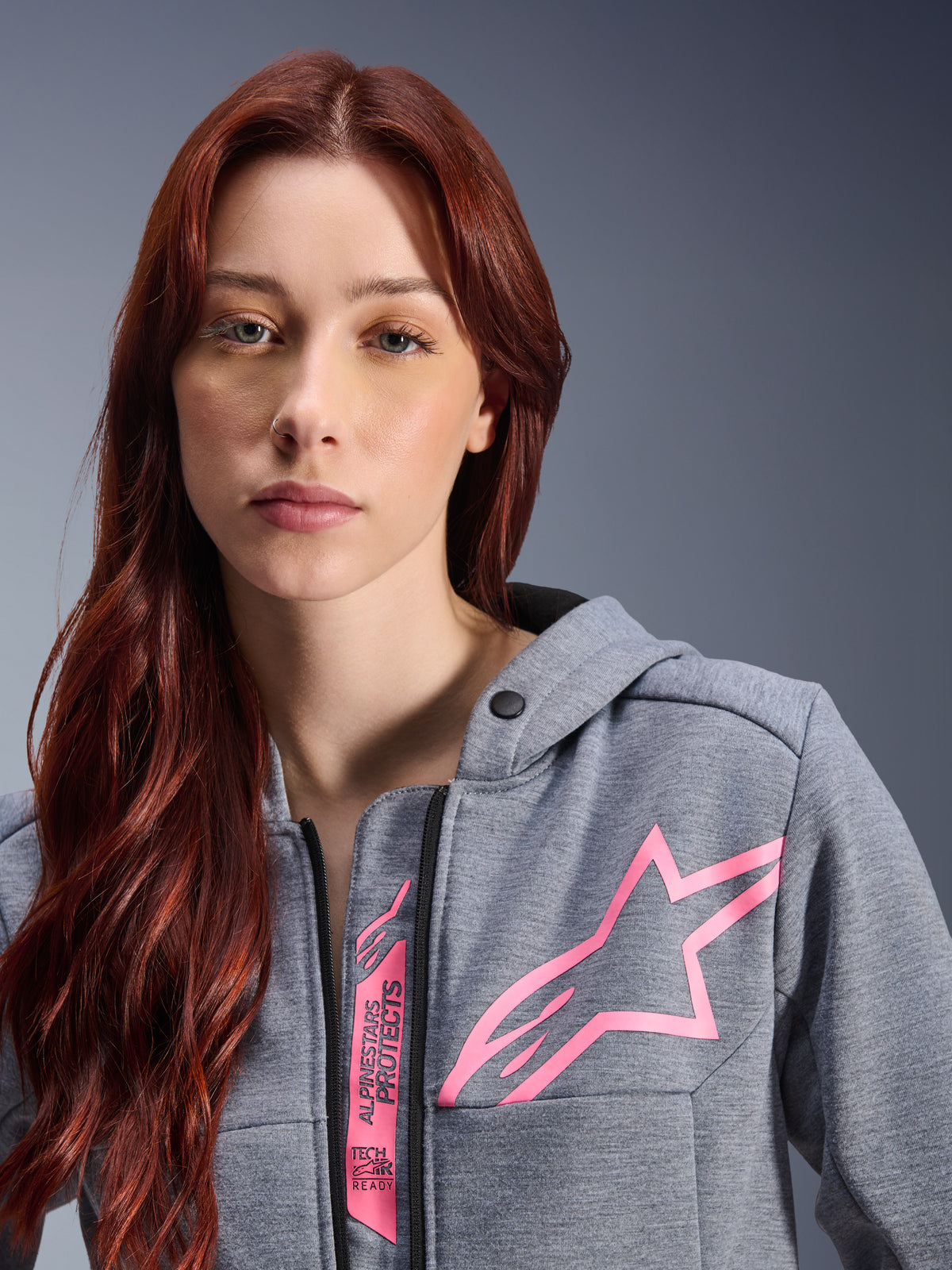 Woman wearing Alpinestars Stella Chrome V2 Sport Hoodie, Gray Heather Pink Fluo, close-up shot showing the pink Alpinestars star logo and Tech-Air Ready branding, urban motorcycle apparel.