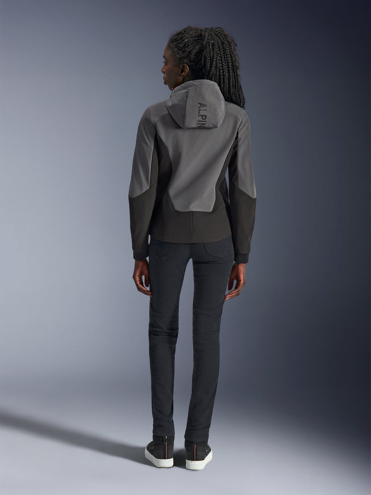 Woman wearing Alpinestars Stella Radium Tech Hoodie, Deep Gray and Black, back view showing 'ALPIN' branding on the hood, paired with slim-fit black pants and black sneakers, urban motorcycle riding apparel