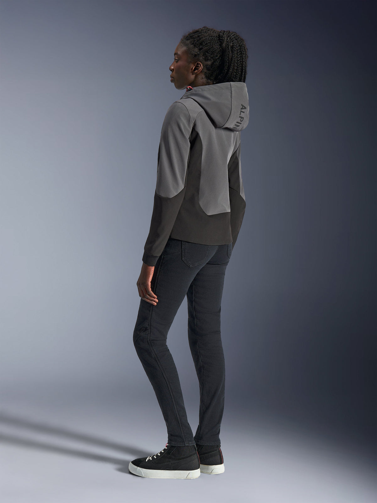 Woman wearing Alpinestars Stella Radium Tech Hoodie in Deep Gray and Black, urban motorcycle apparel, back view showing hood with Alpinestars branding, styled with black slim-fit pants and high-top sneakers