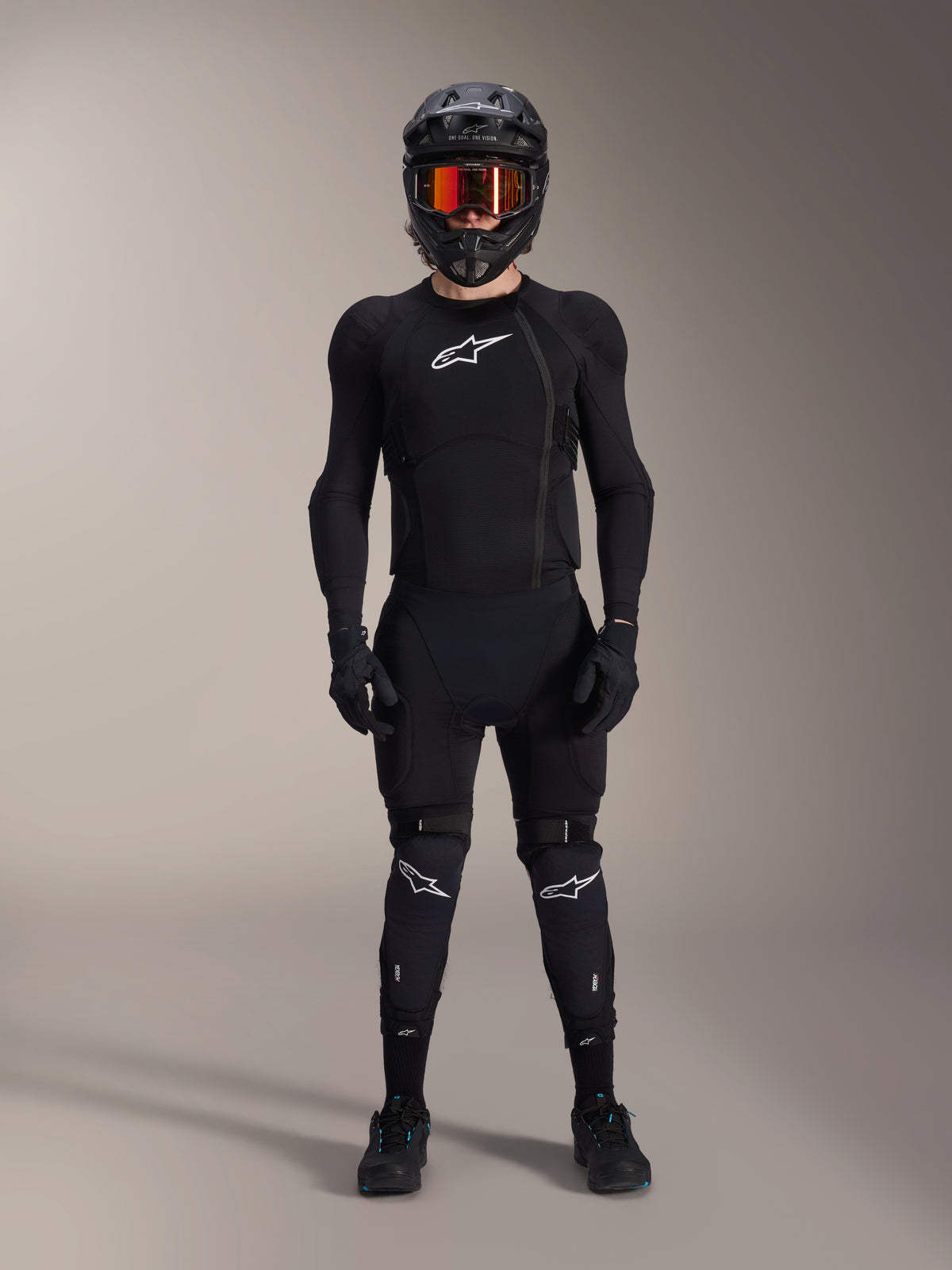 Mountain bike rider wearing complete protective gear, Alpinestars A-Impact Plasma Protection Jacket in Black and White, featuring a white chest logo and mesh fabric, worn with a black helmet, red-tinted goggles, and knee guards, coordinated high-performance MTB armor set.