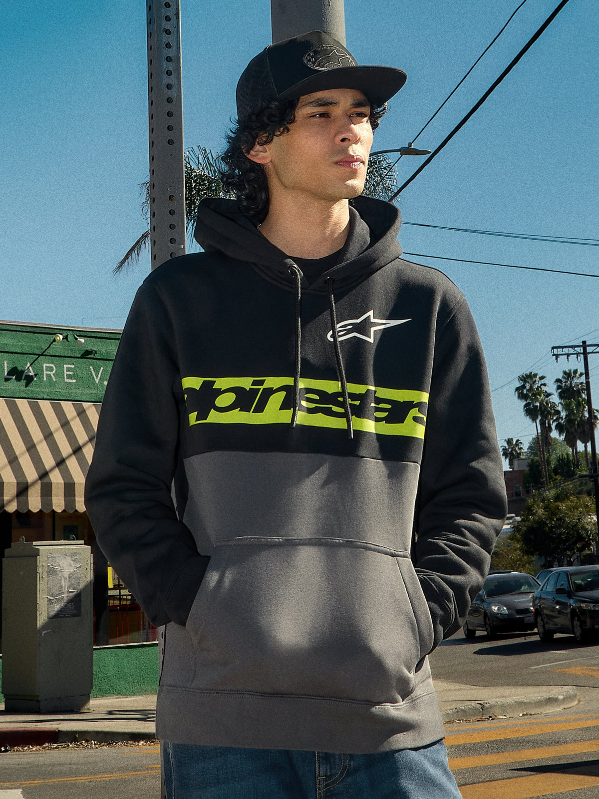 Alpinestars Parallel Hoodie, lifestyle streetwear, color-blocked black and gunmetal design, lime green chest stripe with Alpinestars logo, white Astars logo, drawstring hood, worn by a man in an outdoor urban setting.