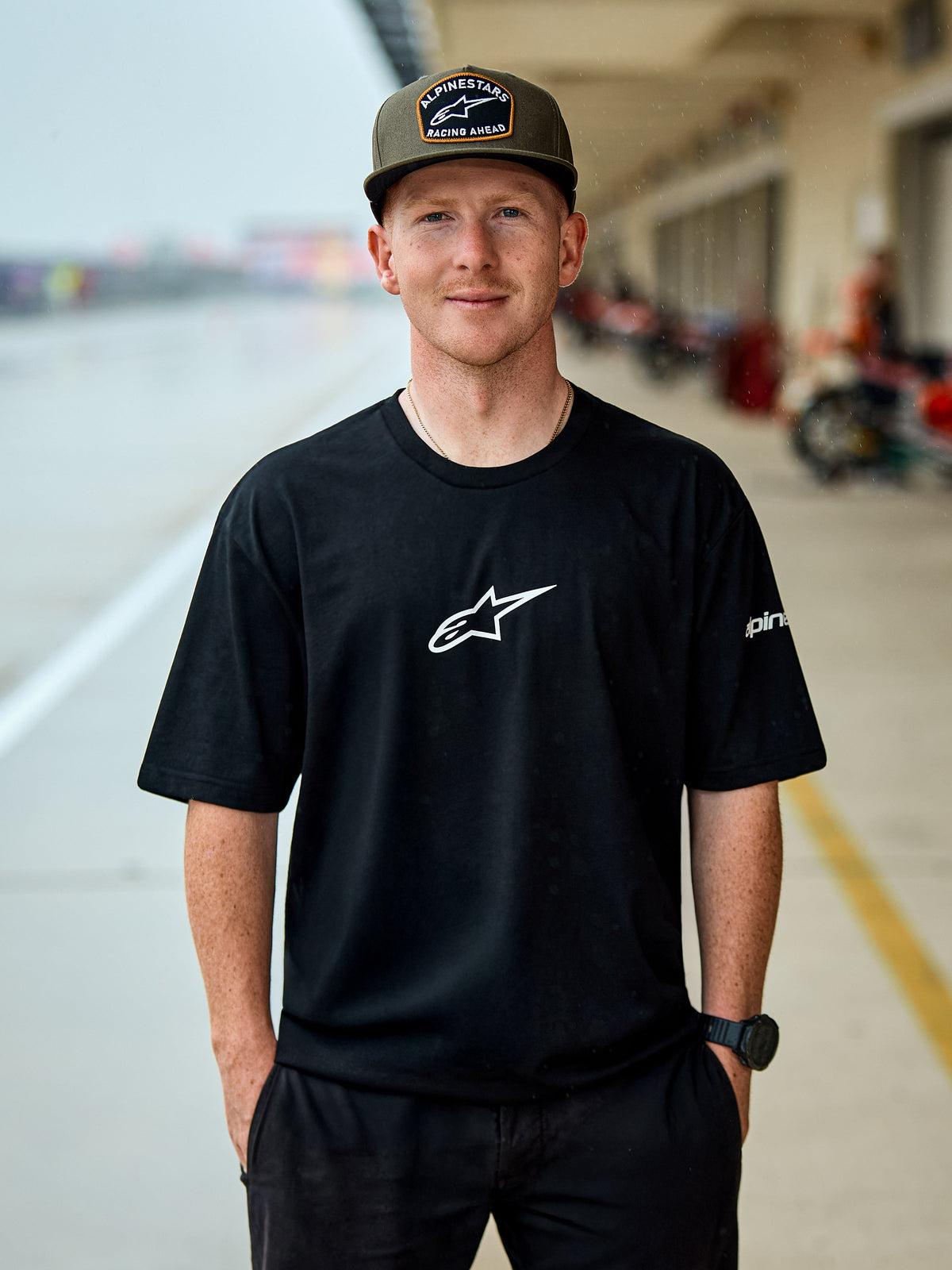 Man wearing Alpinestars Frontal CSF Tee, Short Sleeve T-Shirt, Black, white central Astars logo, olive green Alpinestars cap, casual motorsport-inspired streetwear