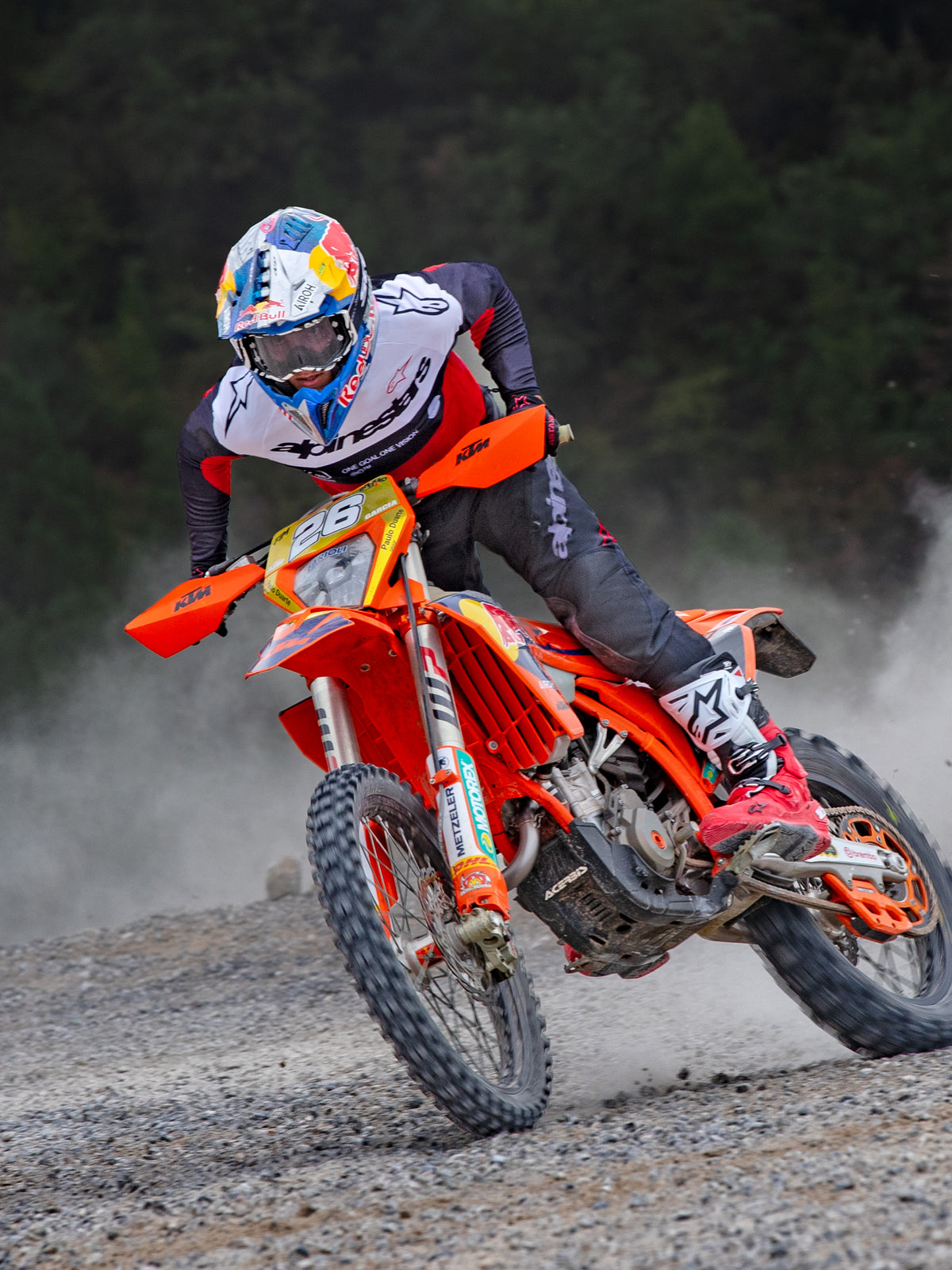 Motocross rider in action on an orange KTM bike, wearing Alpinestars Pro-Dura gear including a red, white, and black jersey and dark grey pants, red and white boots, and a Red Bull racing helmet, performing a sharp turn on a gravel trail