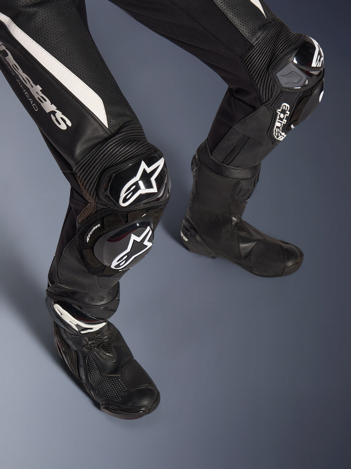 Detail of Alpinestars Stella GP-R7 2-Piece Leather Suit legs, Black with white accents, featuring GP DFS RR knee armor and sliders with Alpinestars branding, paired with black racing boots, engineered for track performance