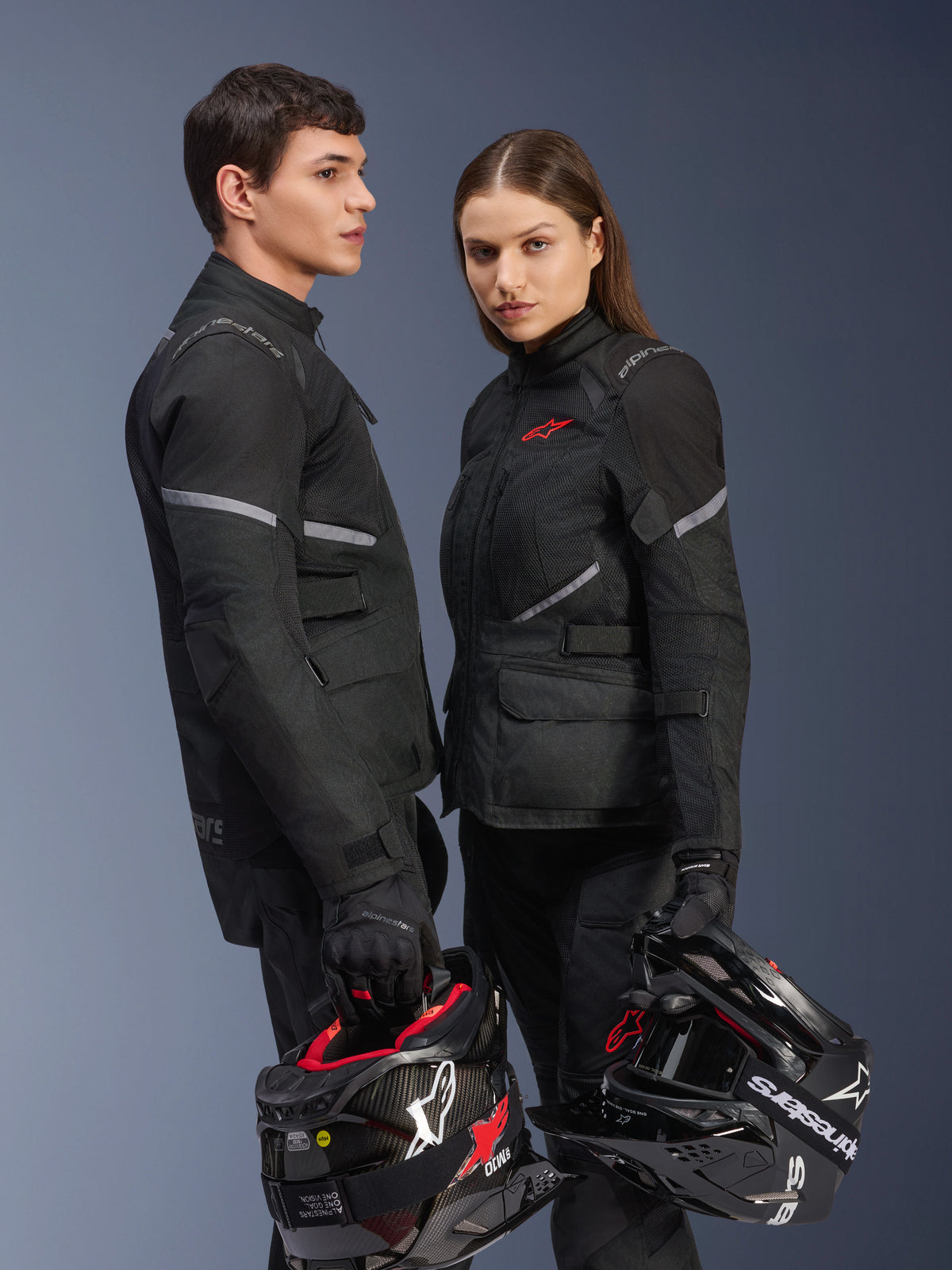 Male and female riders in Alpinestars motorcycle gear, featuring the Stella Andes Air Drystar® Jacket in Black, matching textile riding pants and gloves, both holding black and white helmets, studio shot showing coordinated adventure touring apparel