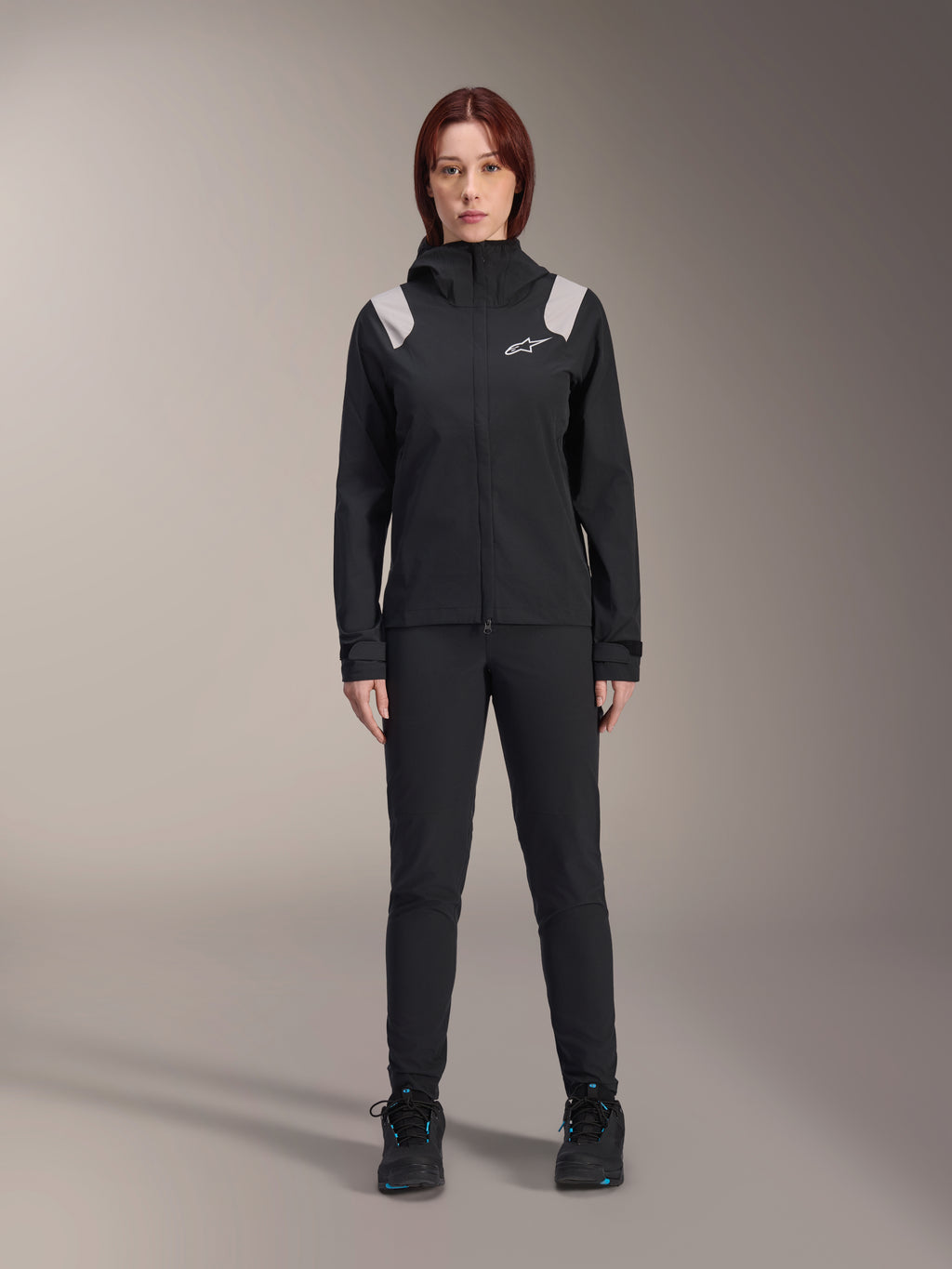Alpinestars Stella A-Dura Rain Jacket, Women's MTB Rain Jacket, Black with light gray shoulder accents and white logo, worn with matching black pants and shoes, featuring a hood and adjustable cuffs, designed for mountain bike trail riding