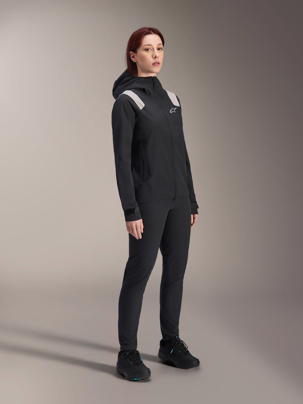 Woman wearing the Alpinestars Stella A-Dura Rain Jacket in Black, 2.5-layer waterproof MTB shell with light grey shoulder accents, matching black pants and cycling shoes, studio portrait for performance cycling gear