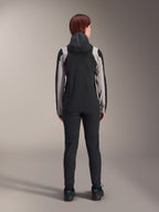 Female rider from behind wearing Alpinestars Stella A-Dura Rain Jacket, Black with light grey panels on the sleeves and shoulders, hood up, worn with black pants and cycling shoes, technical mountain bike rain gear