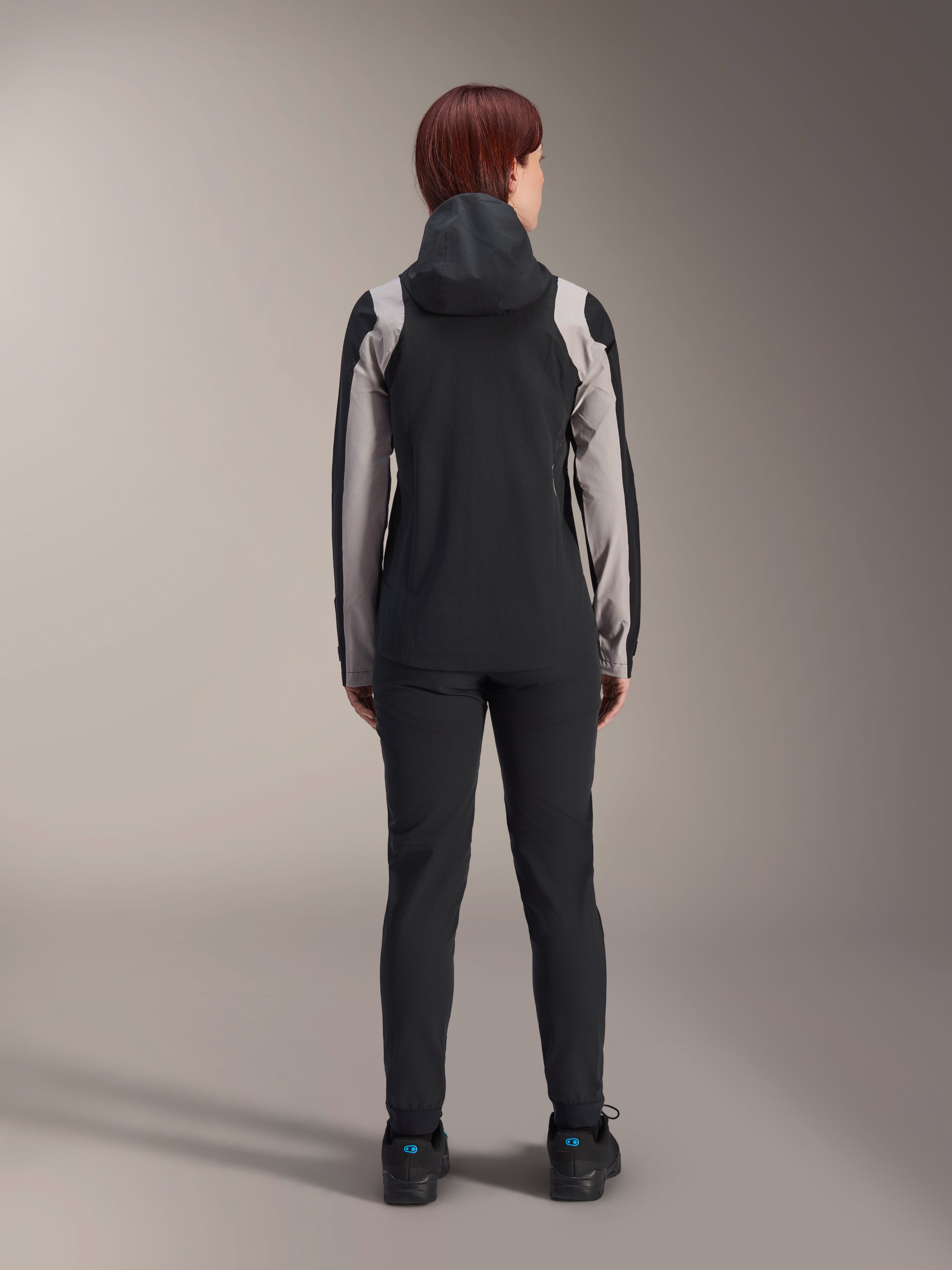 Female rider from behind wearing Alpinestars Stella A-Dura Rain Jacket, Black with light grey panels on the sleeves and shoulders, hood up, worn with black pants and cycling shoes, technical mountain bike rain gear