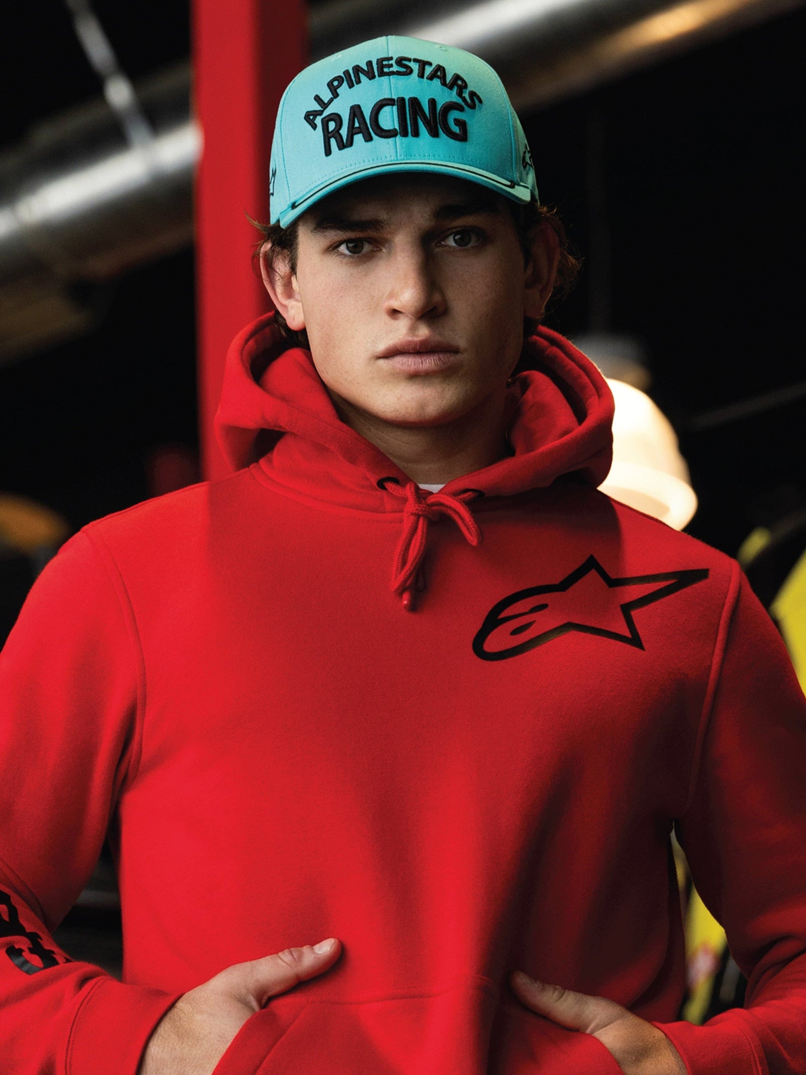 Alpinestars Luminary Hat, Aqua Blue curved bill snapback, black 3D embroidery with 'ALPINESTARS RACING' text, worn with a bright red Alpinestars hoodie, casual motorsport lifestyle streetwear