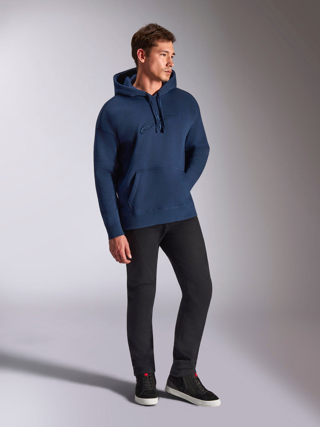 Alpinestars Emergent Oversized Hoodie, Navy Blue, worn by a male model with black pants and sneakers, featuring 3D embroidered Astars logo, kangaroo pocket, and drawstring hood, casual motorsport-inspired streetwear.