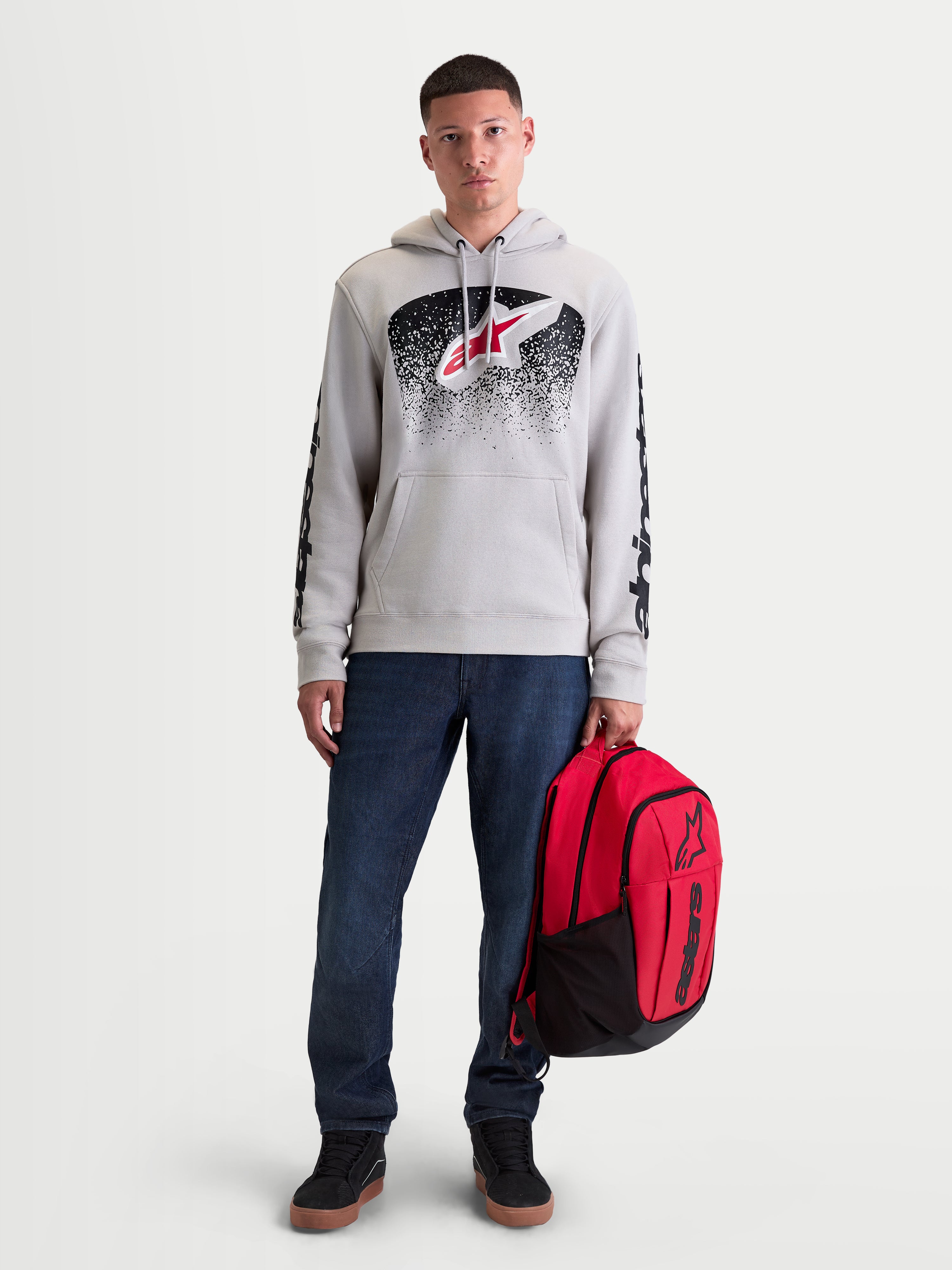 Alpinestars Obsessed Hoodie, Men's Sportswear Pullover Fleece, Cement Gray with Red and Black MX-inspired gradient graphics. This casual motorsport lifestyle hoodie features sleeve branding and is shown on a male model holding a red Alpinestars backpack.