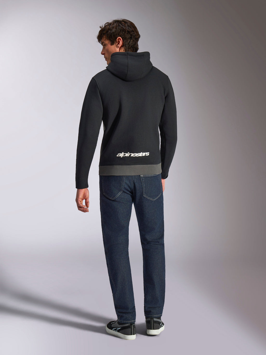 Alpinestars Parallel Hoodie, rear view of a man wearing a black and gunmetal color-blocked pullover fleece, white Alpinestars word logo on the lower back, paired with dark blue jeans, casual motorsport-inspired streetwear