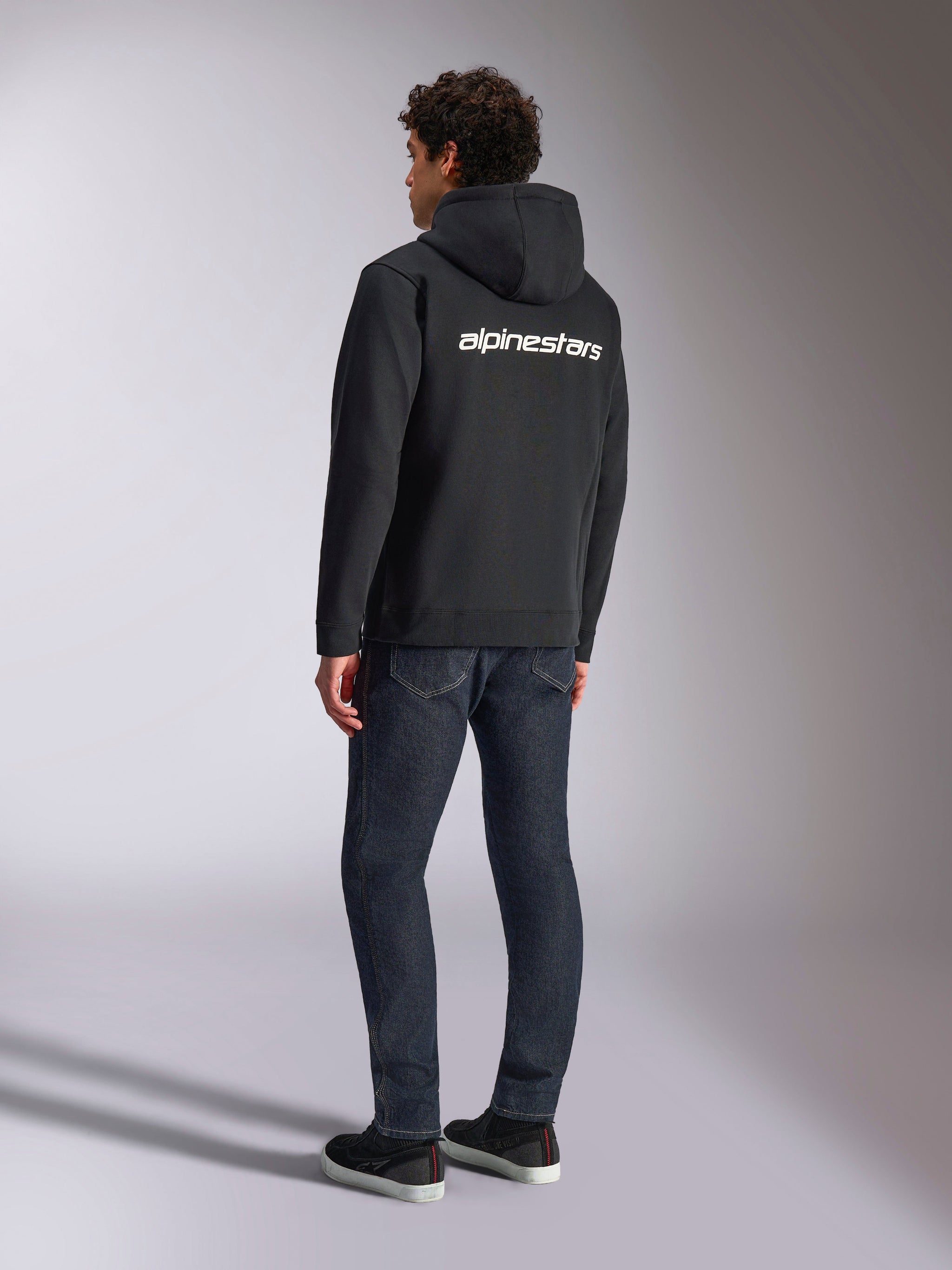 Alpinestars Transverse Hoodie, Black zip-front fleece, back view featuring a white Alpinestars word logo across the shoulders, worn with dark blue jeans and black sneakers, casual motorsport-inspired streetwear