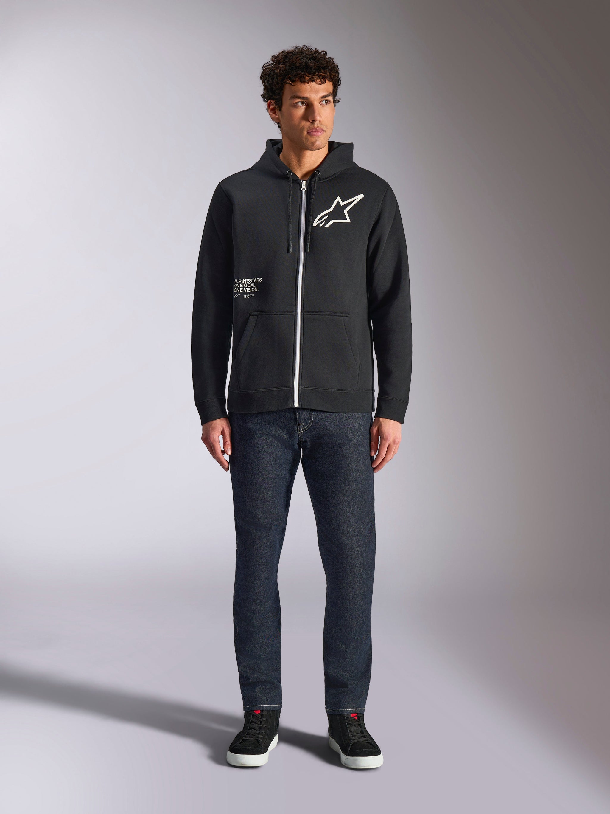 Alpinestars Transverse Hoodie, casual zip-front fleece, Black, white Astars logo on chest and 'One Goal' print on front, drawstring hood, paired with dark jeans and black sneakers, lifestyle motorsport streetwear