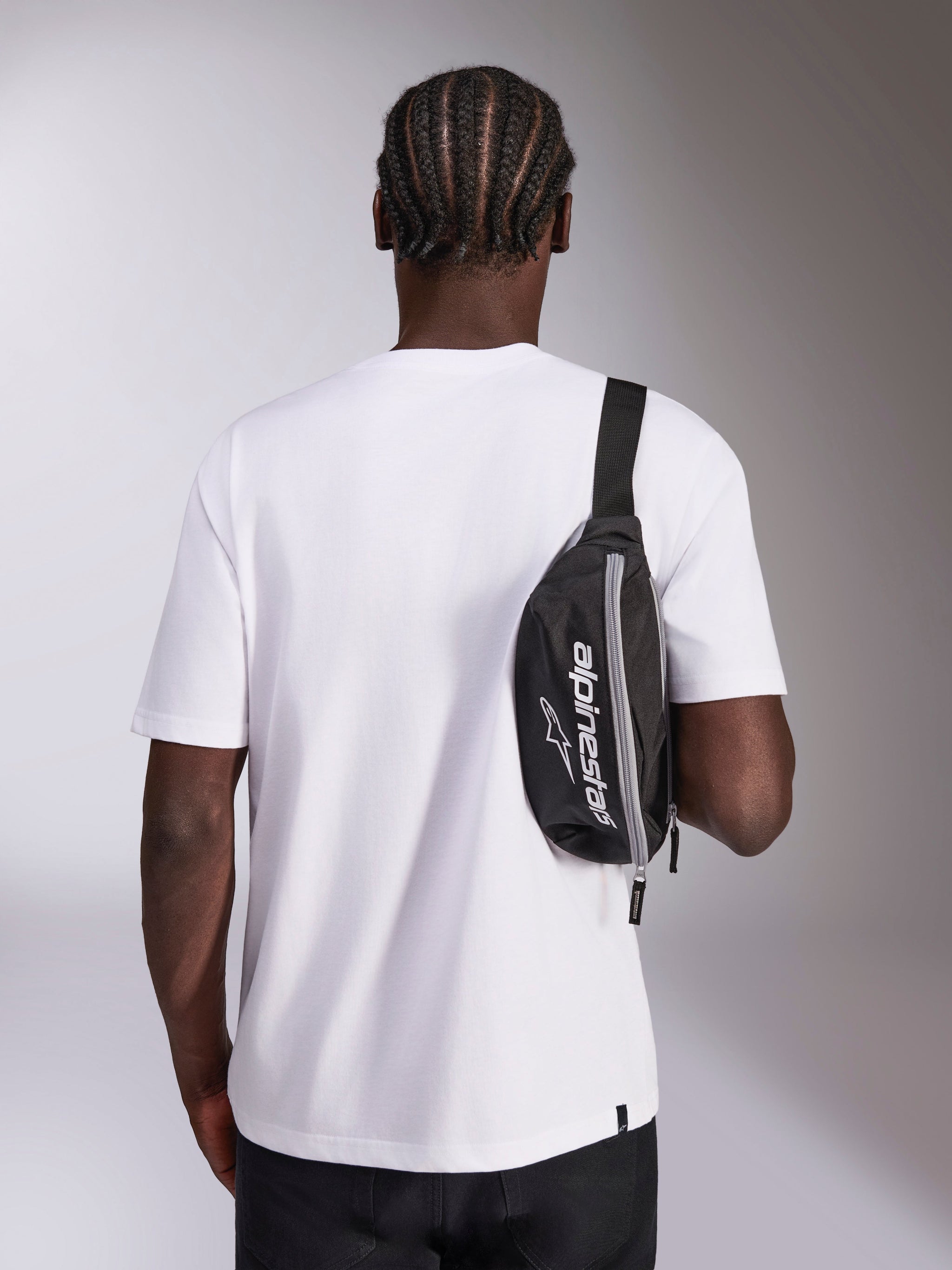 Alpinestars Only Linear Hip Bag, black with white Alpinestars logo branding, worn as a crossbody bag over a white t-shirt, casual motorsport-inspired streetwear accessory
