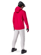 Alpinestars A-Dura Elite Waterproof Jacket, Red Fluo, three-quarter back view, worn with light gray pants and black MTB helmet, features a hood and reflective accents, high-performance cycling rain jacket for trail riding