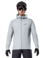 Alpinestars A-Dura Thermal Jacket in Titanium, light gray hooded cycling jacket with a red star logo on the chest, worn by a rider with a black MTB helmet, sunglasses, and gloves, insulated gear for mountain biking
