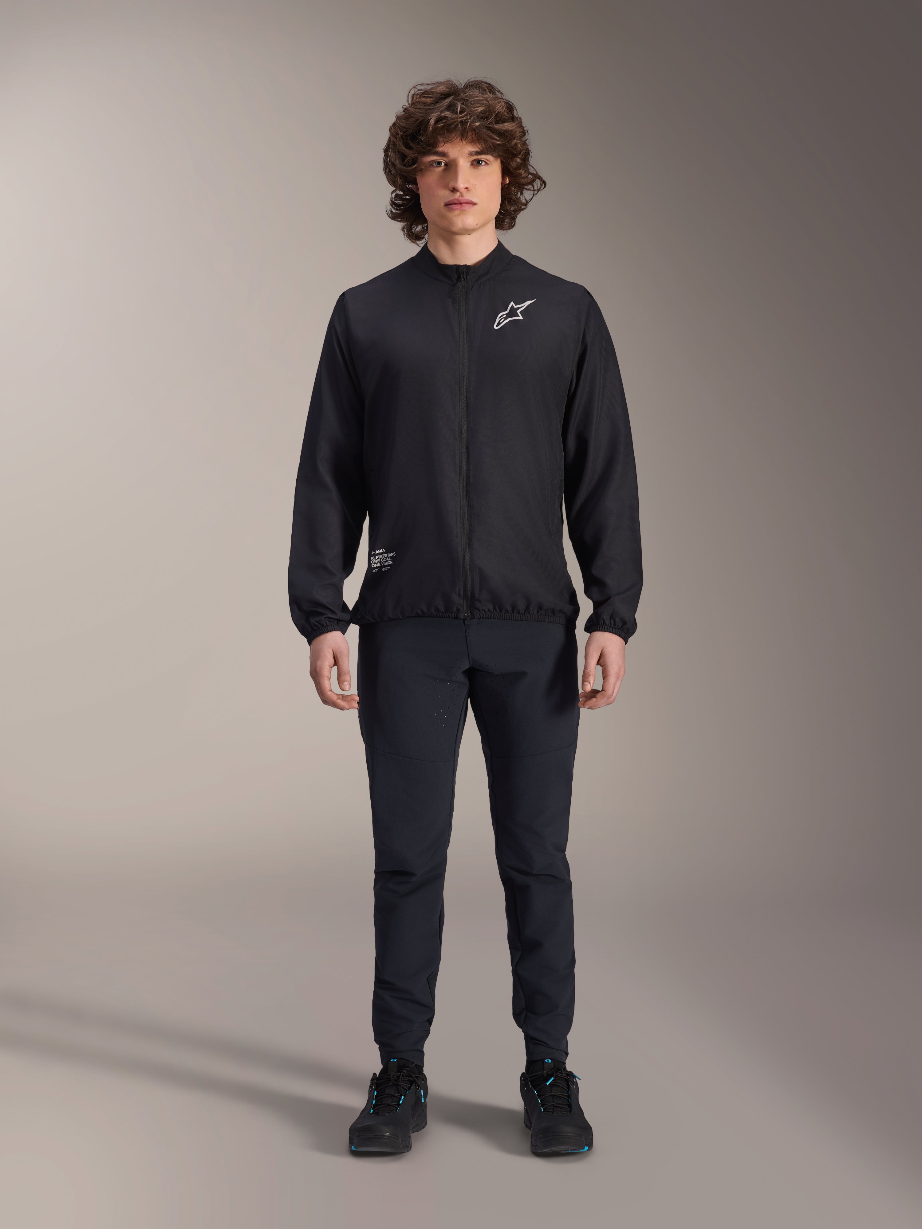 Model wearing Alpinestars A-Aria Windbreaker Jacket in Black, featuring a full-zip front, mock neck, and white Astars logo on the chest, paired with black technical pants and black shoes, performance cycling apparel for mountain biking and outdoor use.