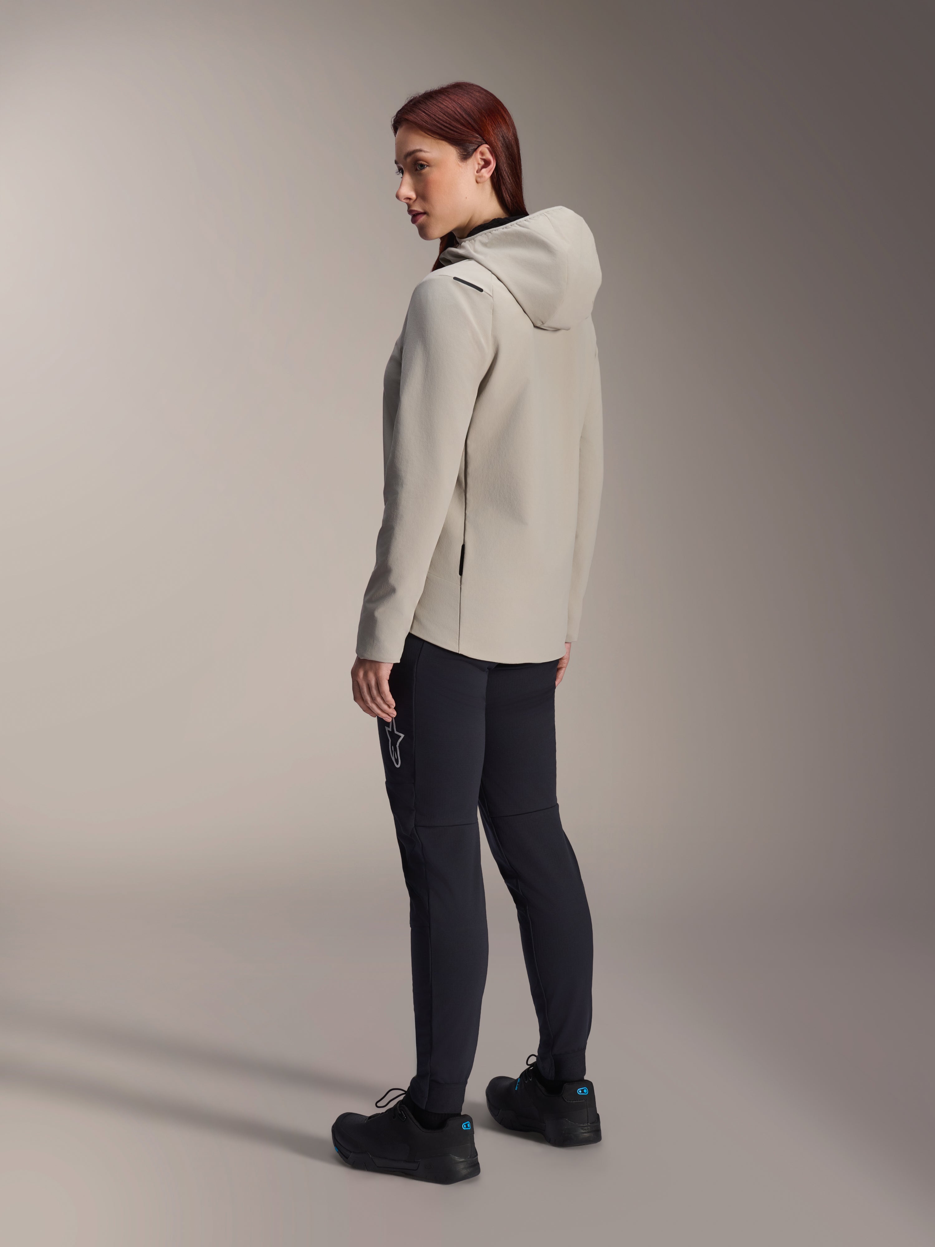 Alpinestars Stella A-Dura Thermal Jacket in Titanium Gray, three-quarter rear view of a woman wearing the insulated hooded cycling jacket with black MTB pants, studio shot showing the regular fit and pocket detail.