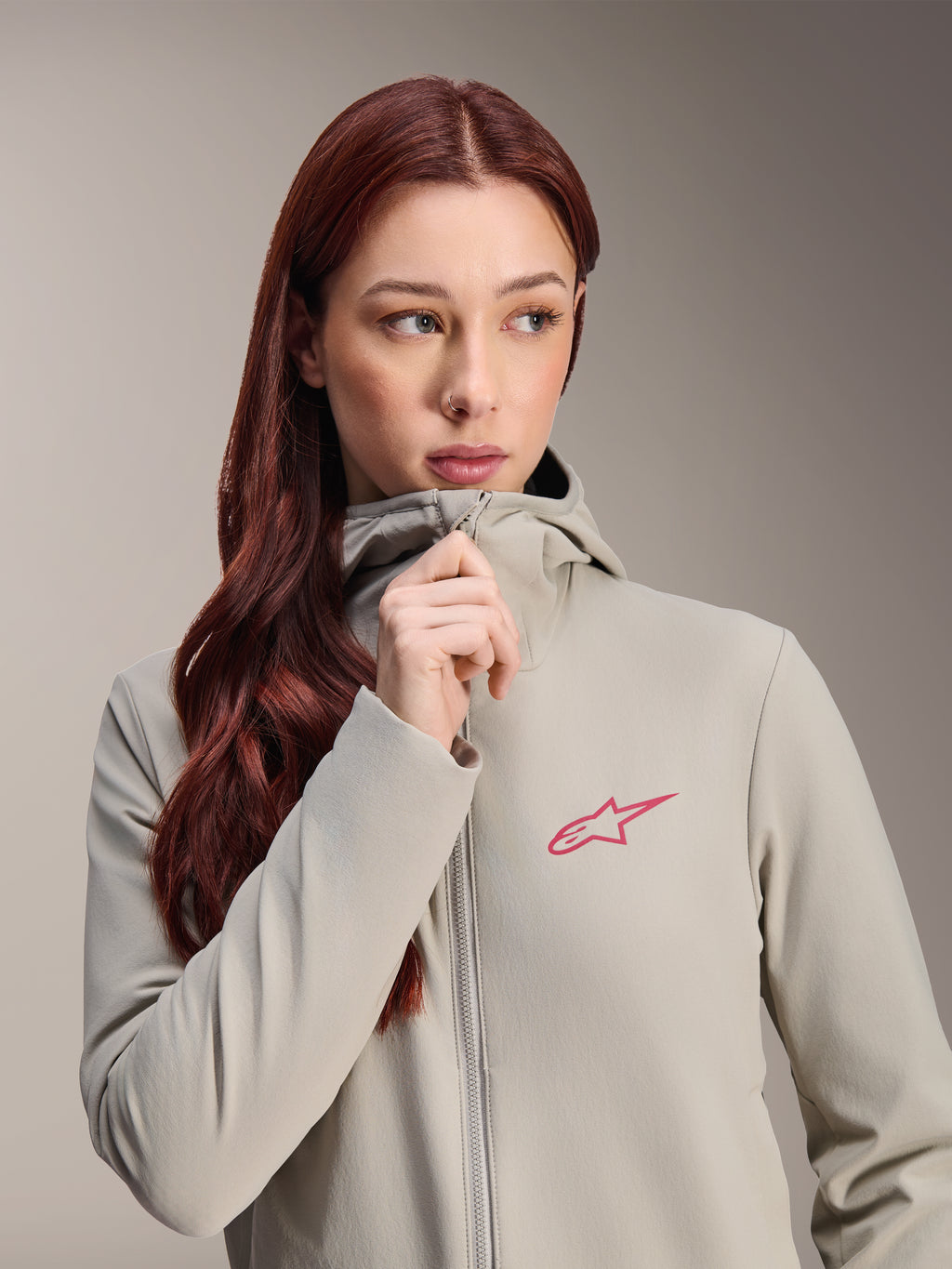 Alpinestars Woman Stella A-Dura Thermal Jacket, Titanium gray with a pink Alpinestars star logo, model with long red hair wearing and zipping up the hooded jacket, lightweight insulated cycling apparel for cold weather.